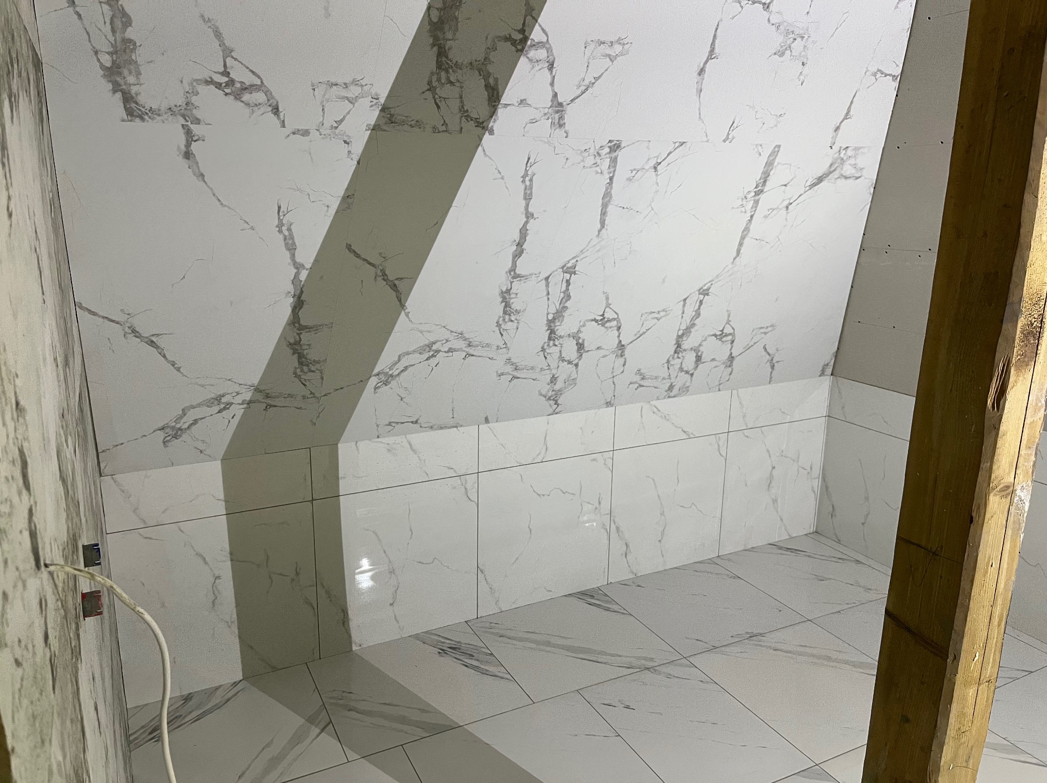 Białe płytki imitujące marmur na ścianach i podłodze poddasza, widoczne fragmenty instalacji elektrycznej i drewnianej konstrukcji, ukończone wykończenie łazienki.