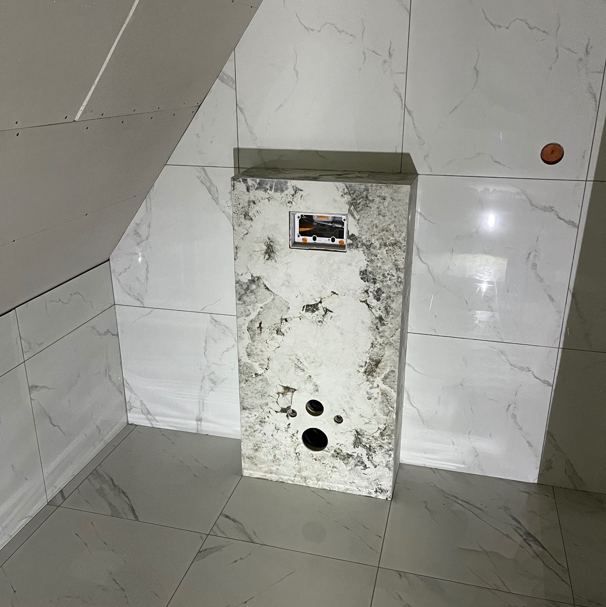 Obudowa geberitu wykończona płytkami imitującymi marmur w łazience na poddaszu. Widoczne płytki na ścianach i podłodze. Częściowo widoczna płyta karton-gips na suficie.