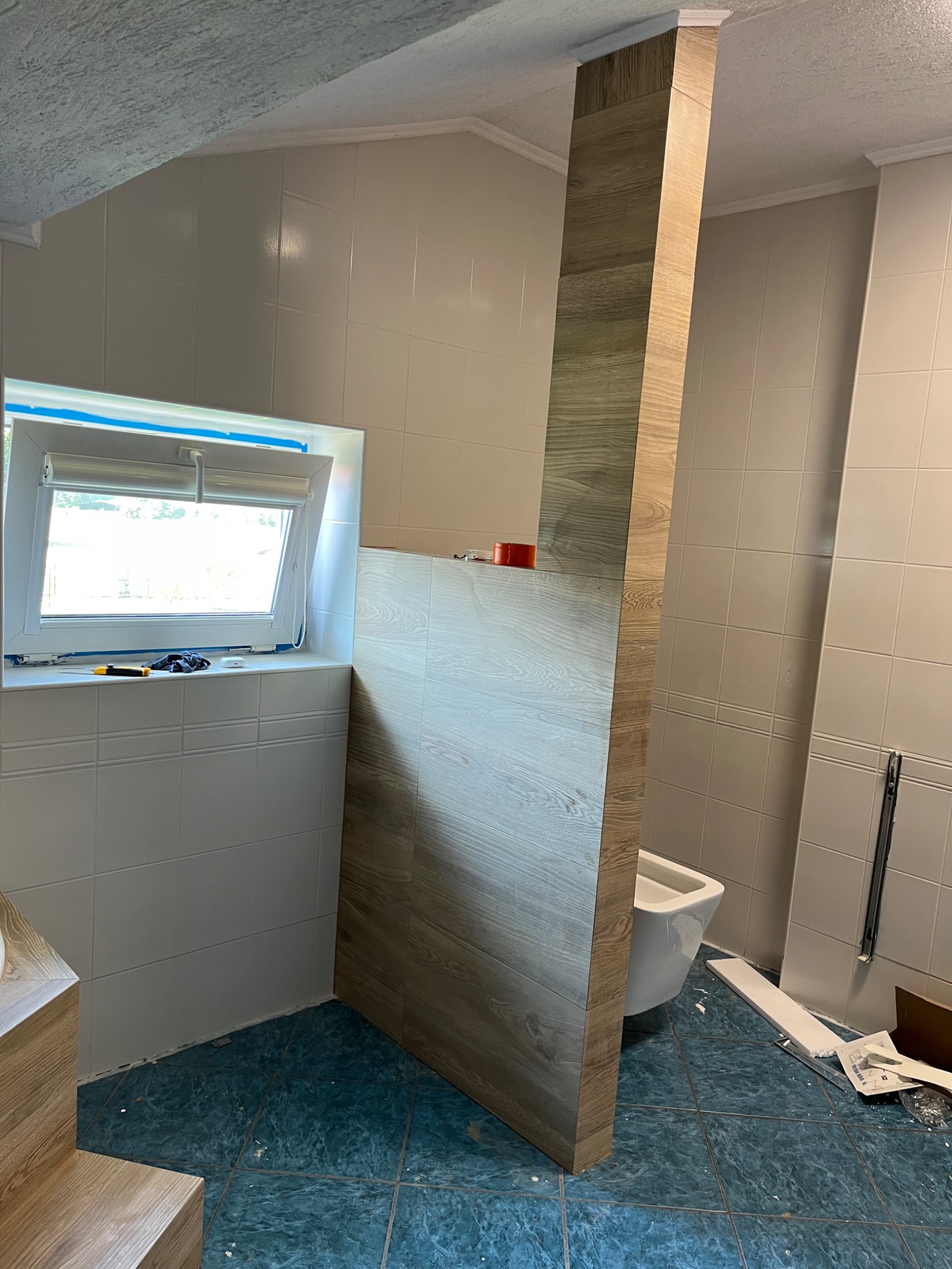 Wnętrze łazienki w trakcie prac: ułożone płytki ścienne i podłogowe, pionowa obudowa z płytek imitujących drewno, okno z narzędziami na parapecie.