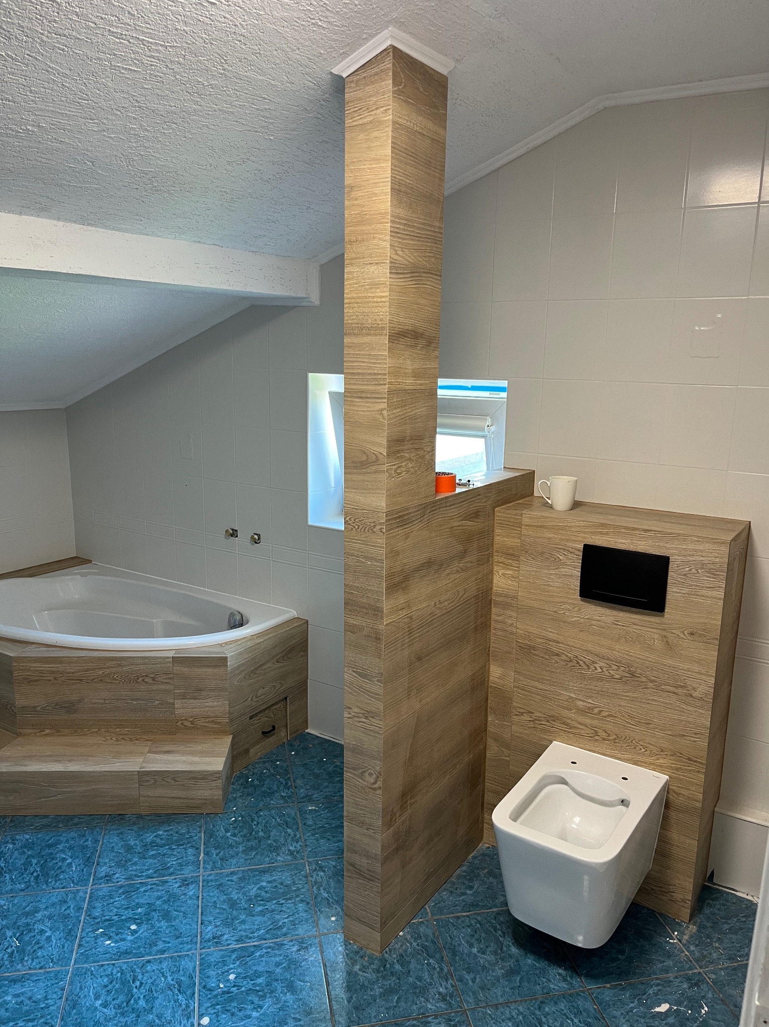 Wykończona łazienka na poddaszu z wanną obudowaną drewnopodobnymi płytkami, toaletą podwieszaną i niebieską podłogą. Widok na okno i białe płytki ścienne.