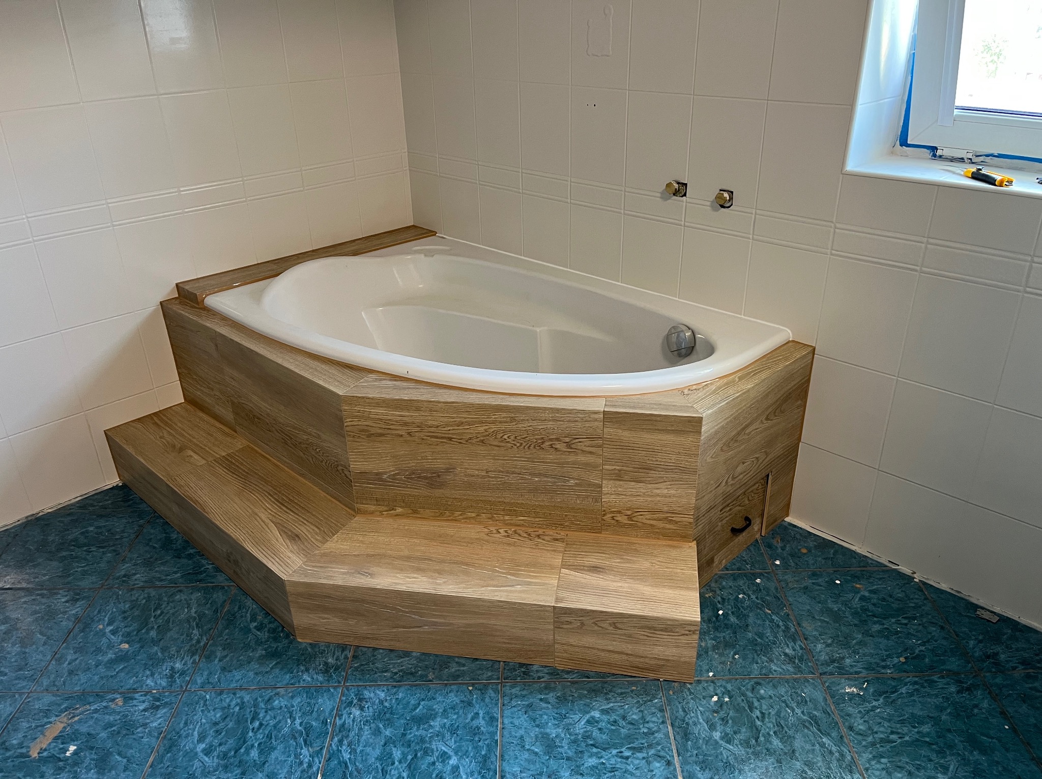 Biała wanna z obudową imitującą drewno, umieszczona na podwyższeniu ze schodkami w łazience z niebieską podłogą i białymi płytkami ściennymi.