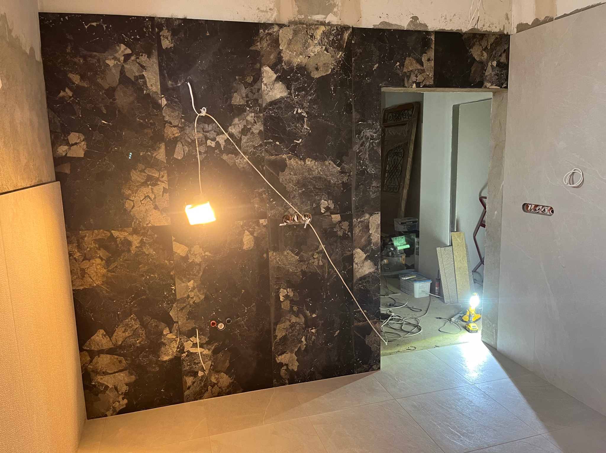Częściowo wykończona ściana z ciemnych, marmurkowych płytek w łazience, widoczne przewody elektryczne i narzędzia, podłoga wyłożona jasnymi płytkami.