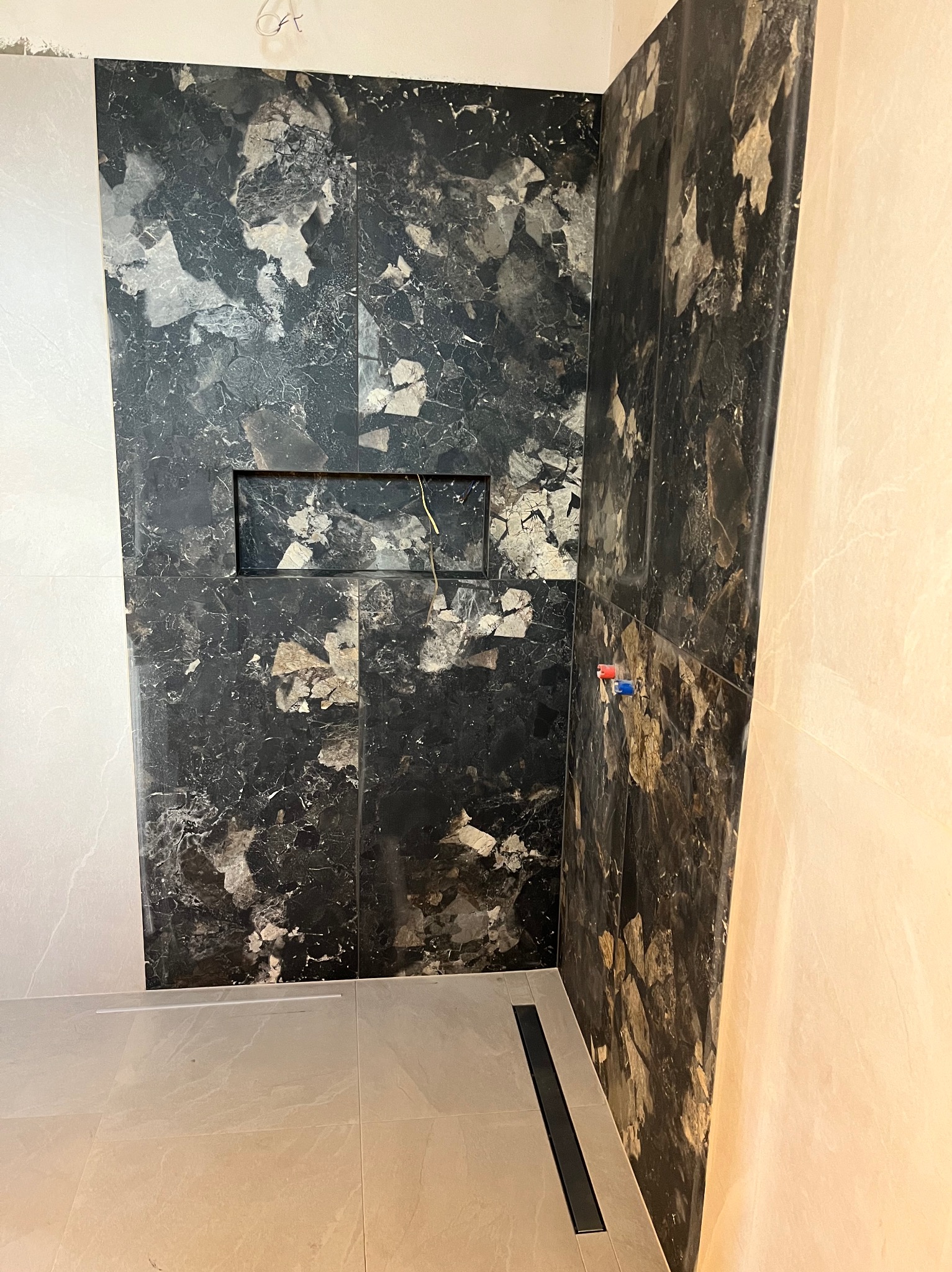 Wykończona łazienka z czarnymi płytkami imitującymi marmur na ścianie prysznicowej z wnęką i odpływem liniowym w podłodze. Widoczne przyłącza wodne.