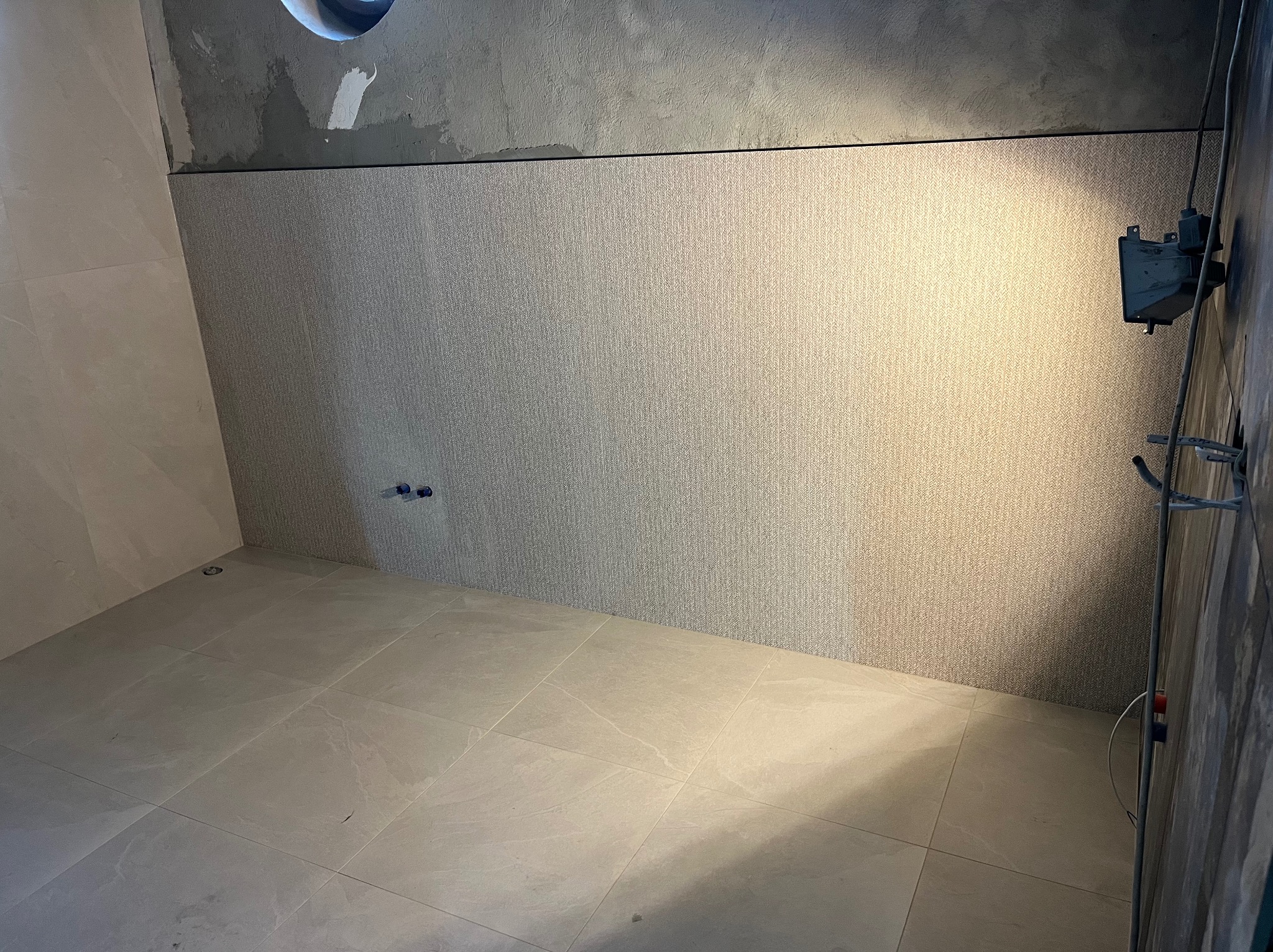 Wykończona łazienka z beżowymi płytkami na podłodze i ścianie do połowy wysokości. Nad płytkami surowy beton. Widoczne przyłącza wodne i instalacja prysznicowa.