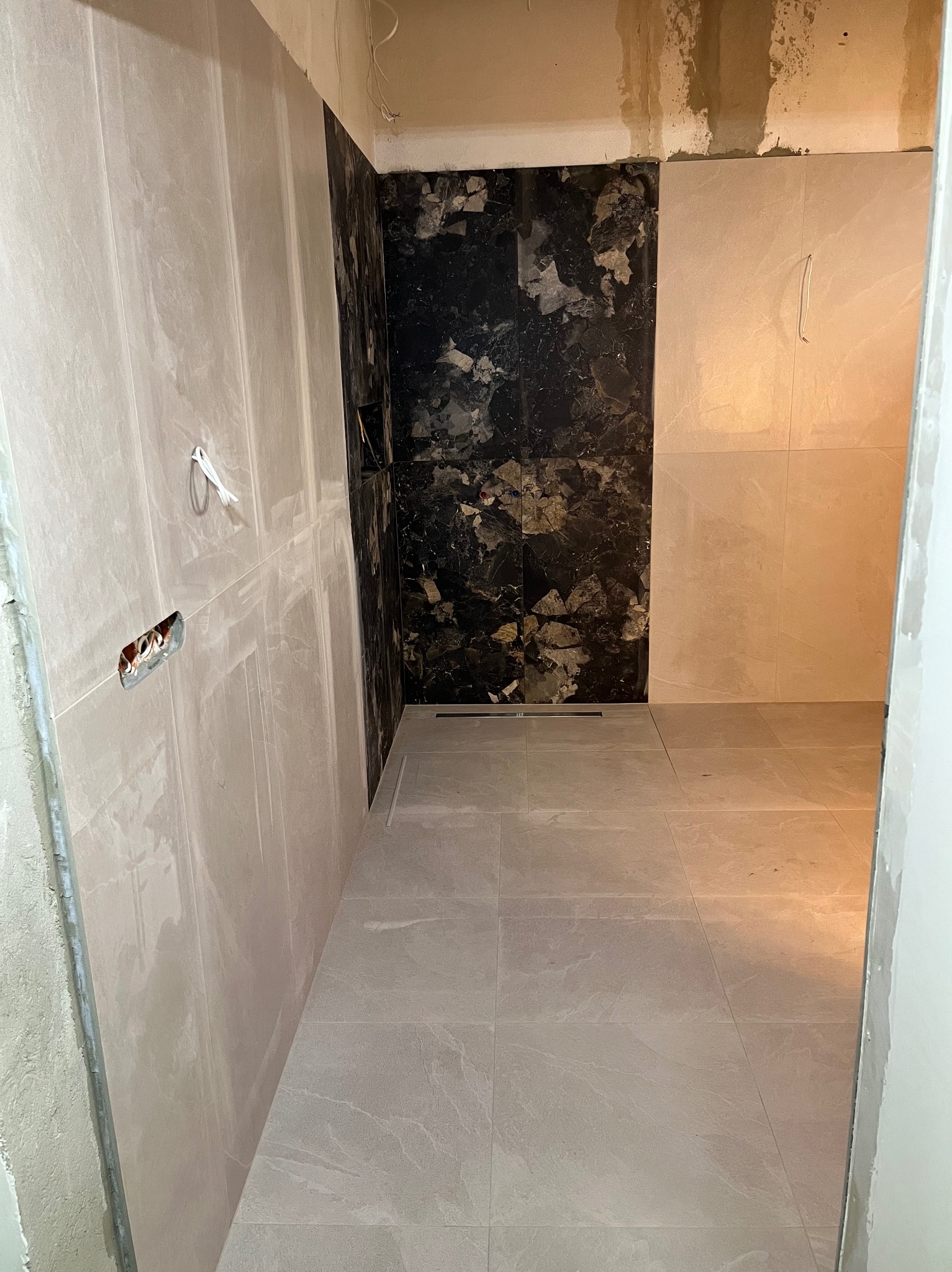 Wnętrze łazienki w trakcie wykańczania: ułożone płytki podłogowe i ścienne w odcieniach beżu i czerni, widoczne gniazdka elektryczne, surowy fragment ściany.