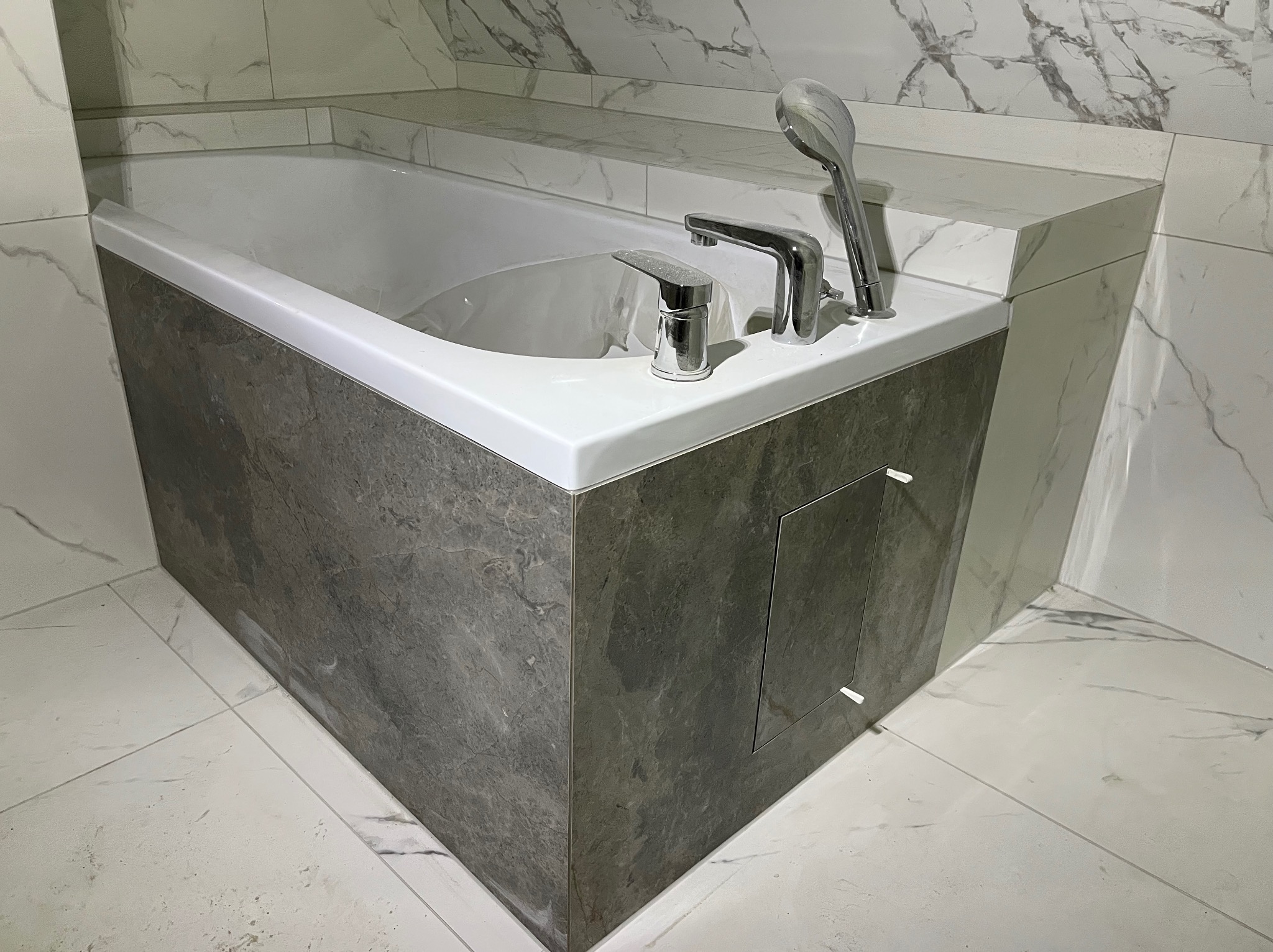 Nowoczesna obudowa wanny z szarych płyt imitujących marmur, biała armatura i płytki w tle. Estetyczne wykończenie łazienki z ukrytą klapą rewizyjną.