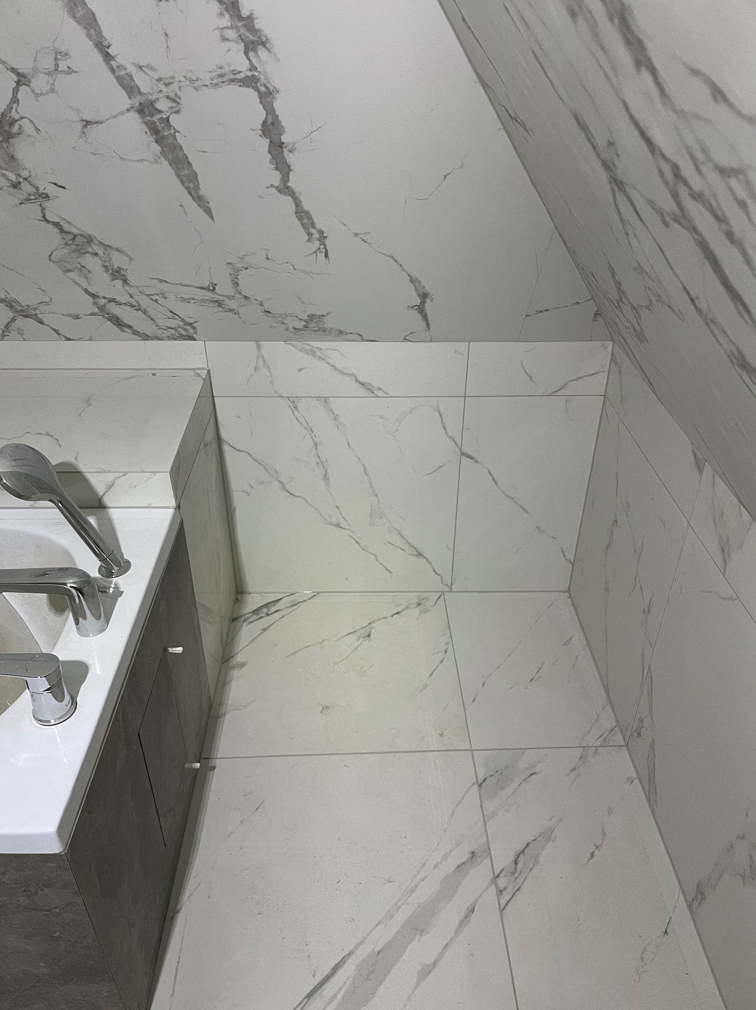 Wykończona łazienka z białymi płytkami imitującymi marmur na podłodze, ścianach i skosach. Widoczna umywalka z szarą szafką i chromowaną armaturą.