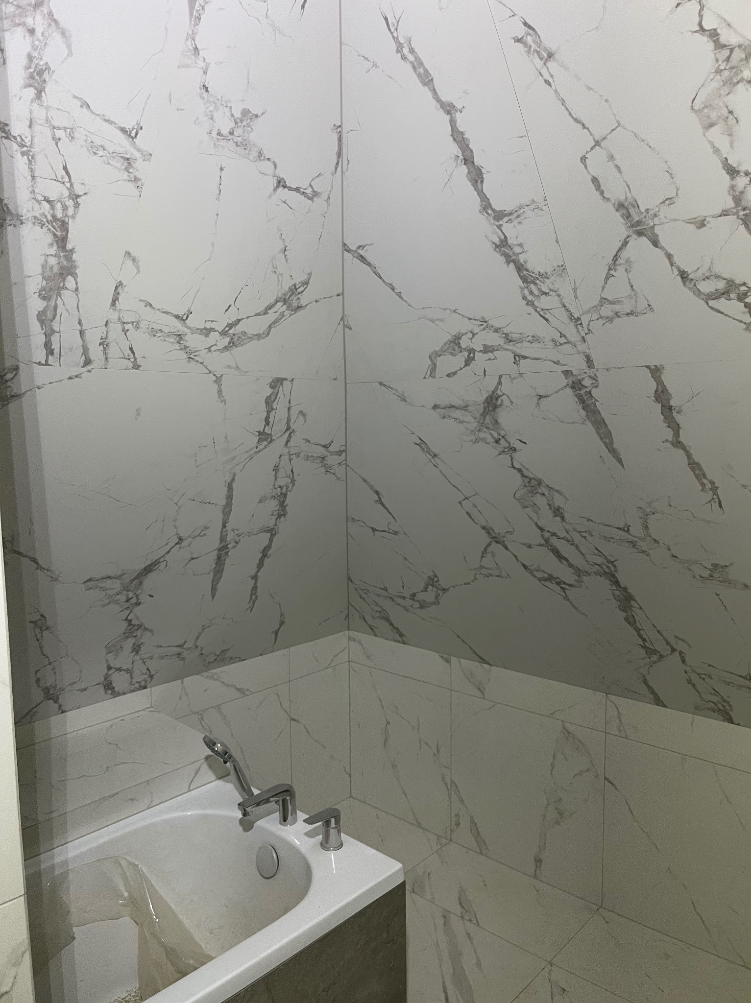 Wykończona łazienka z wanną, ściany i podłoga wyłożone białymi płytkami imitującymi marmur z szarymi żyłami. Bateria wannowa chromowana.