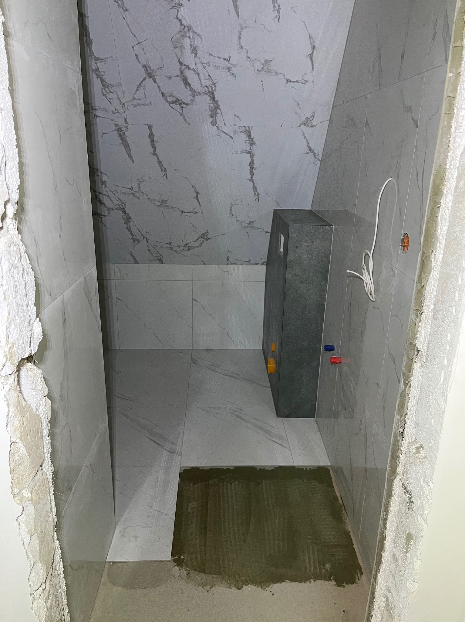 Wnętrze łazienki w trakcie wykończenia: marmurowe płytki na ścianach i podłodze, zabudowa stelaża WC, widoczne instalacje. Surowy stan wykończenia.