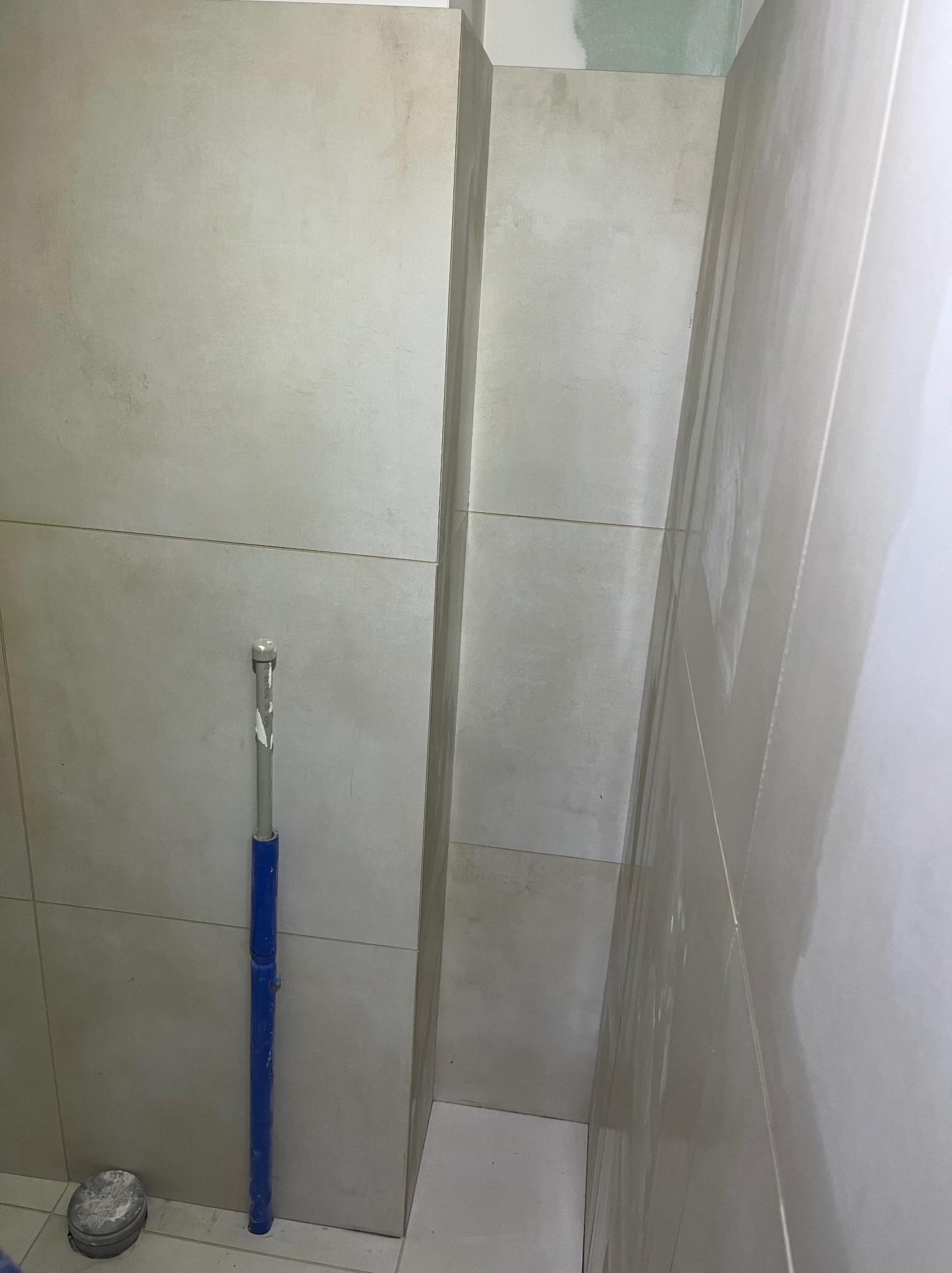 Jasne, matowe płytki w łazience, częściowo ułożone. Widoczny niebieski rozpierak i pojemnik z zaprawą. Świeżo wykończona ściana z płyt karton-gips w górnym rogu.