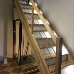 Lunvero - Jasne, drewniane schody z metalowymi balustradami w trakcie montażu, widoczne niedokończone podłoże i narzędzia, oświetlenie LED pod stopniami.