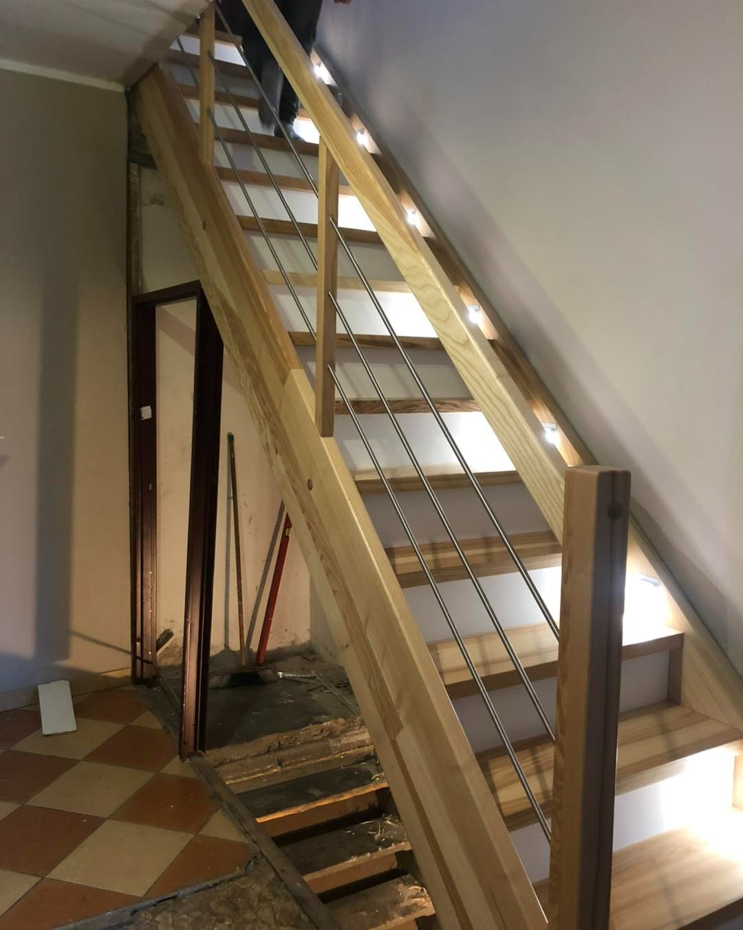 Jasne, drewniane schody z metalowymi balustradami w trakcie montażu, widoczne niedokończone podłoże i narzędzia, oświetlenie LED pod stopniami.
