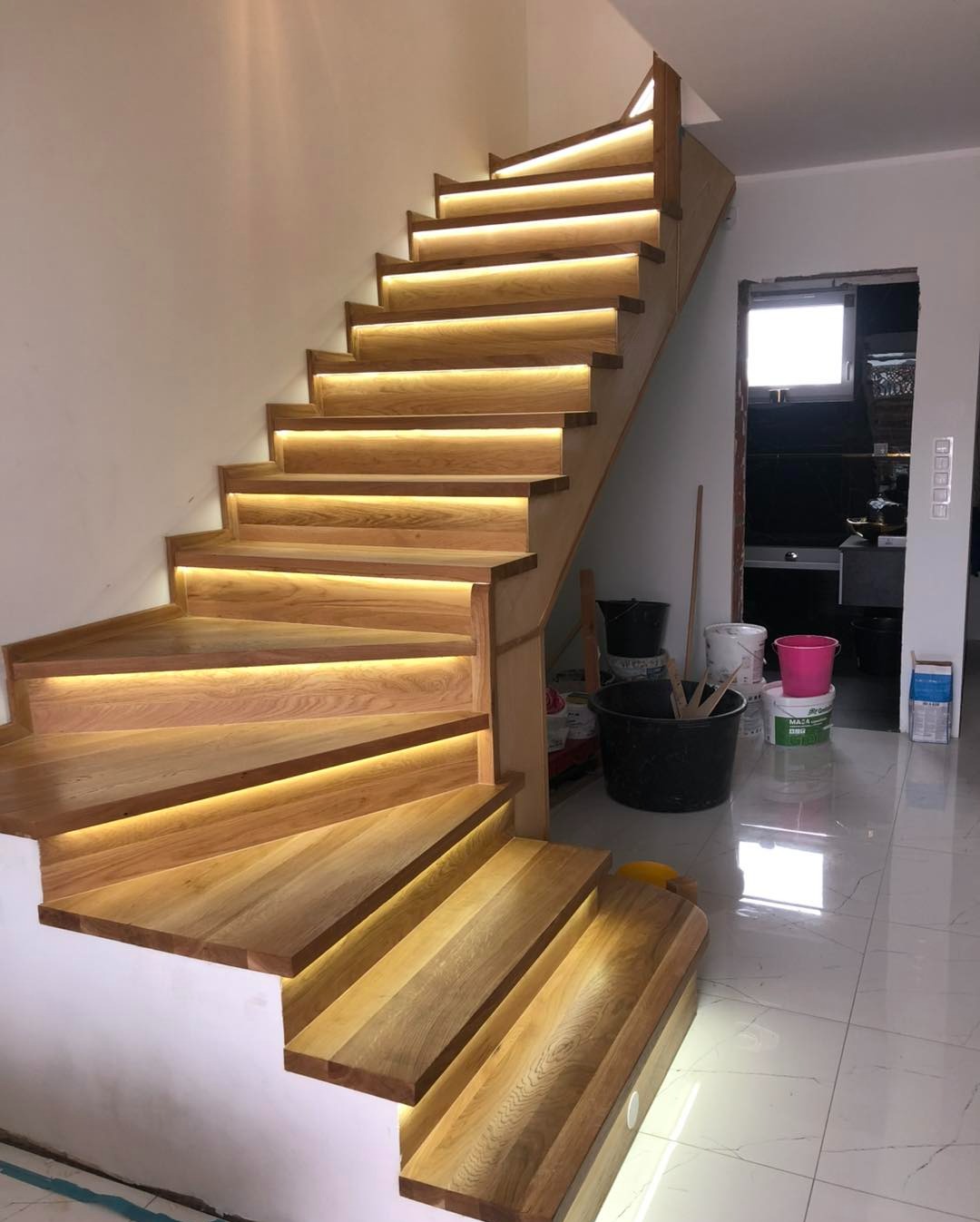 Dębowe schody z podświetleniem LED, widok z dołu. Nowoczesny design, jasne drewno, białe ściany. W tle widoczne wiaderka i narzędzia, co sugeruje prace wykończeniowe.