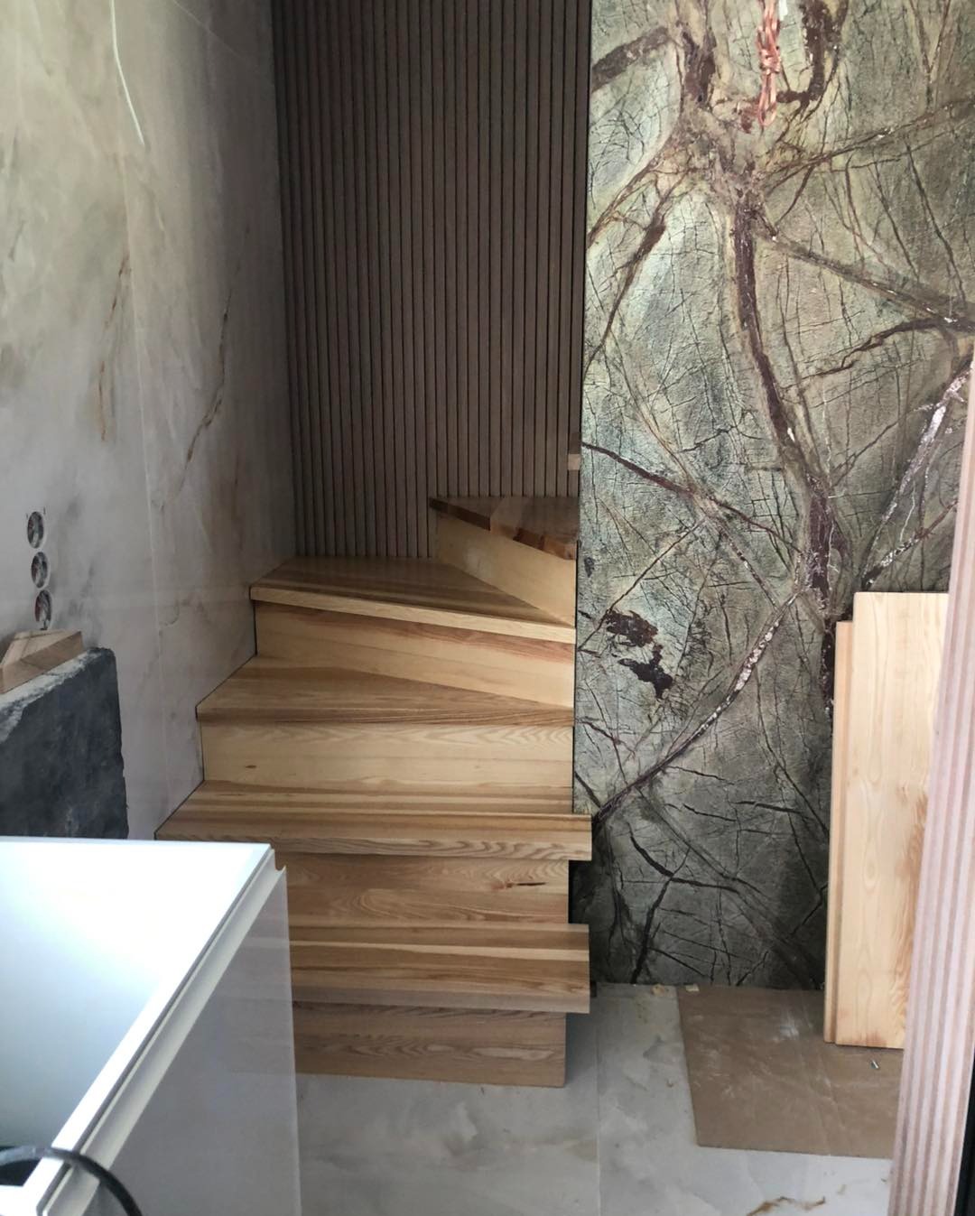 Jasne, drewniane schody w nowoczesnym wnętrzu z marmurowymi ścianami i dekoracyjnym panelem ściennym. Widoczne detale wykończenia i oświetlenie.