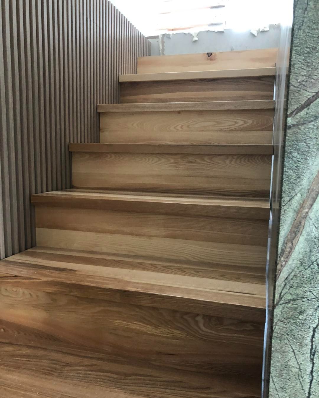 Jasne, drewniane schody z widocznym usłojeniem, z dekoracyjną ścianą z pionowych listew i marmurową okładziną. Ujęcie z dołu, podkreślające naturalny charakter materiałów.