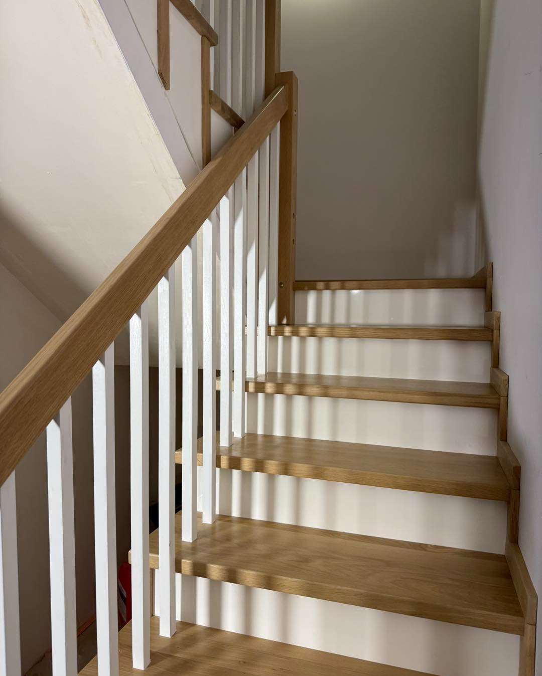 Jasne, drewniane schody z białą balustradą w nowoczesnym wnętrzu. Widoczne stopnie i fragment ściany. Ujęcie z dołu, podkreślające detale wykończenia.