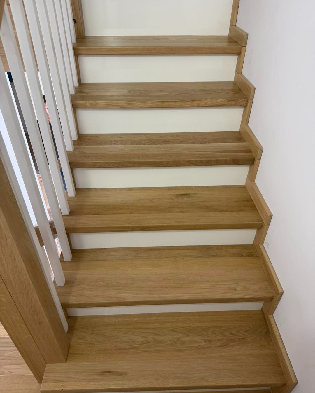 Jasne, dębowe stopnie schodów z białymi podstopnicami i białą balustradą, widziane z góry, z delikatnym oświetleniem podkreślającym strukturę drewna.