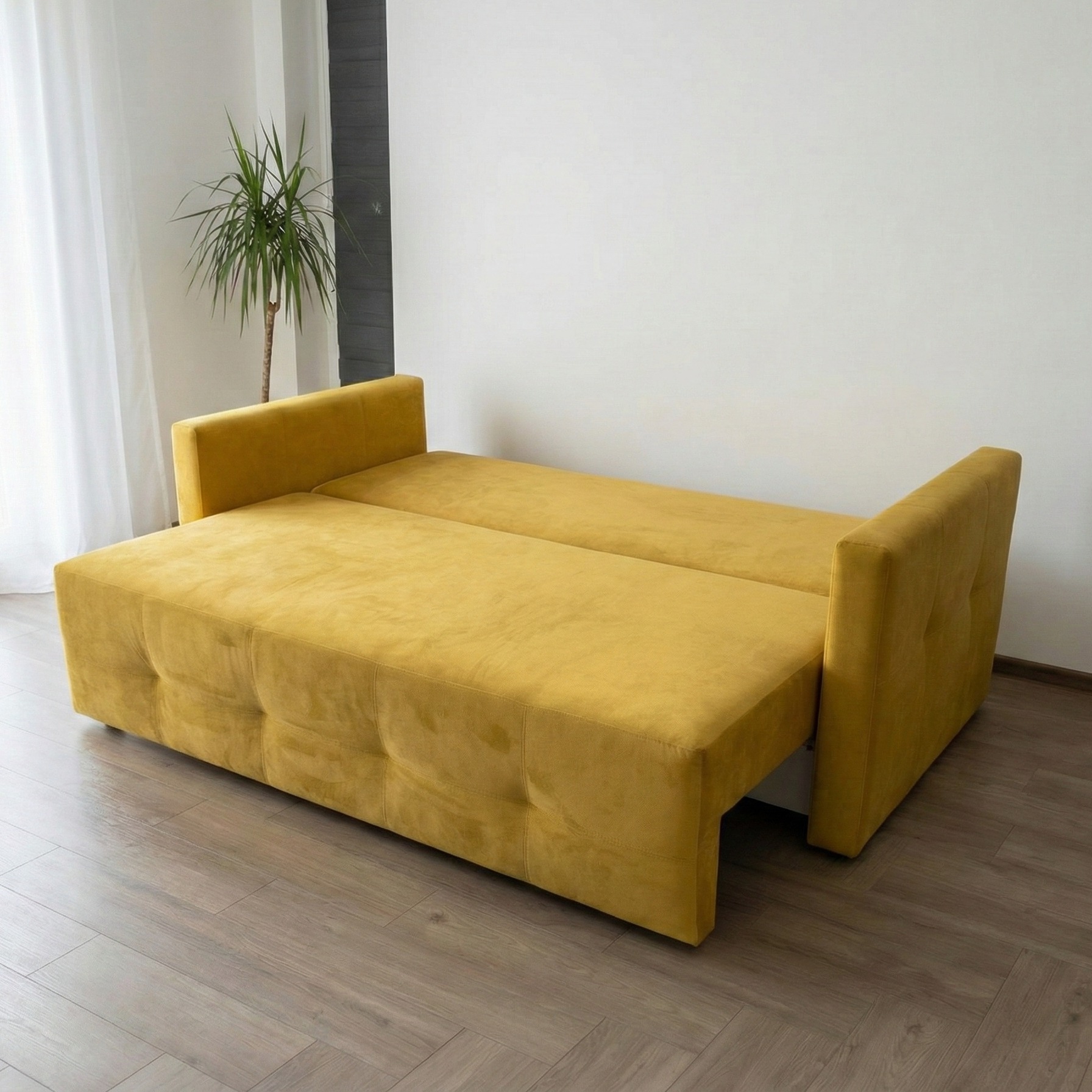 Rozłożona, żółta sofa z podłokietnikami na drewnianej podłodze. W tle biała ściana i roślina doniczkowa. Minimalistyczny wystrój wnętrza.