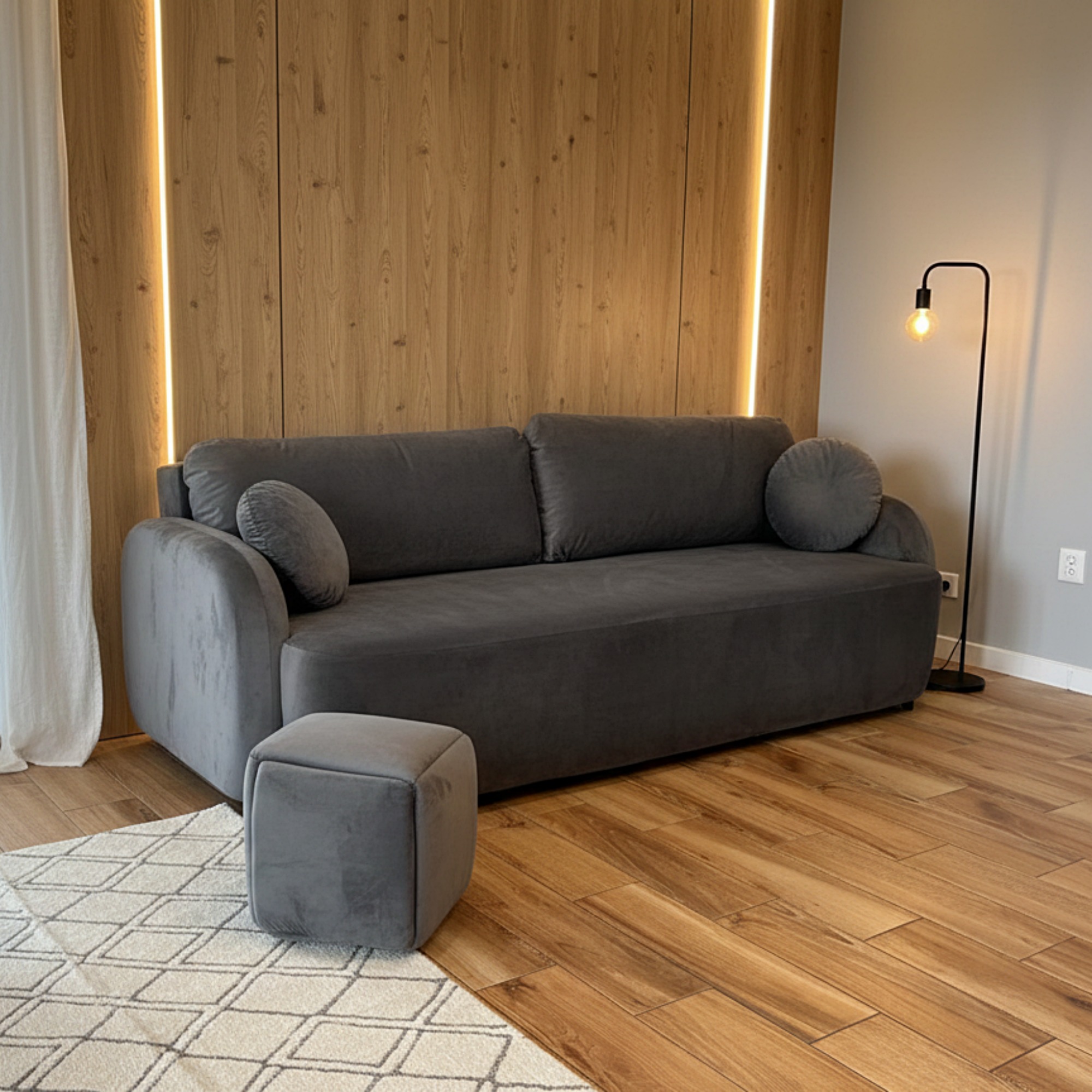 Szara sofa z podnóżkiem na wzorzystym dywanie, na tle drewnianej ściany z oświetleniem LED i lampy podłogowej. Nowoczesny design wnętrza.