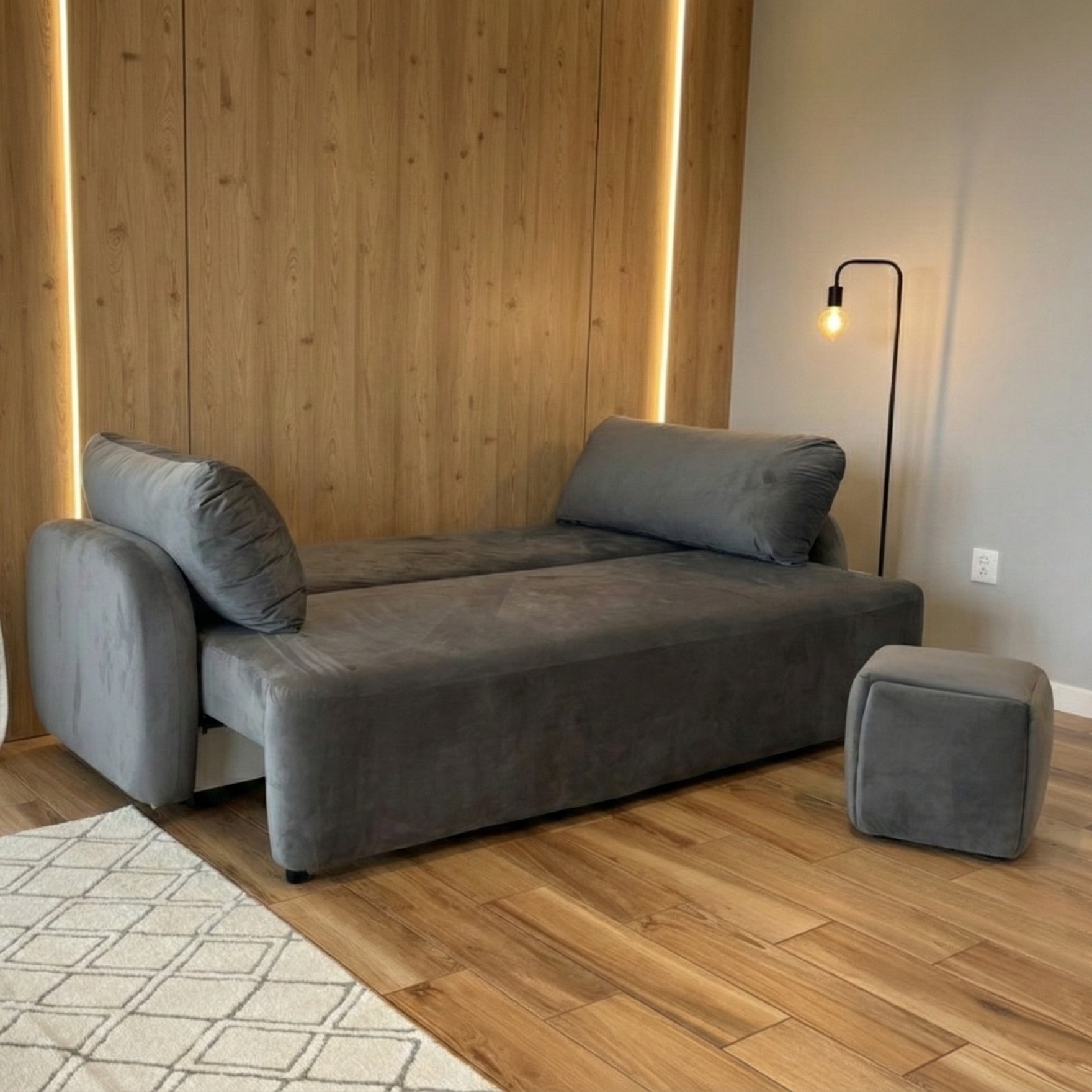 Rozkładana sofa w odcieniach szarości na drewnianej podłodze, z pufą i lampą w tle. Realizacja z Biskupic.
