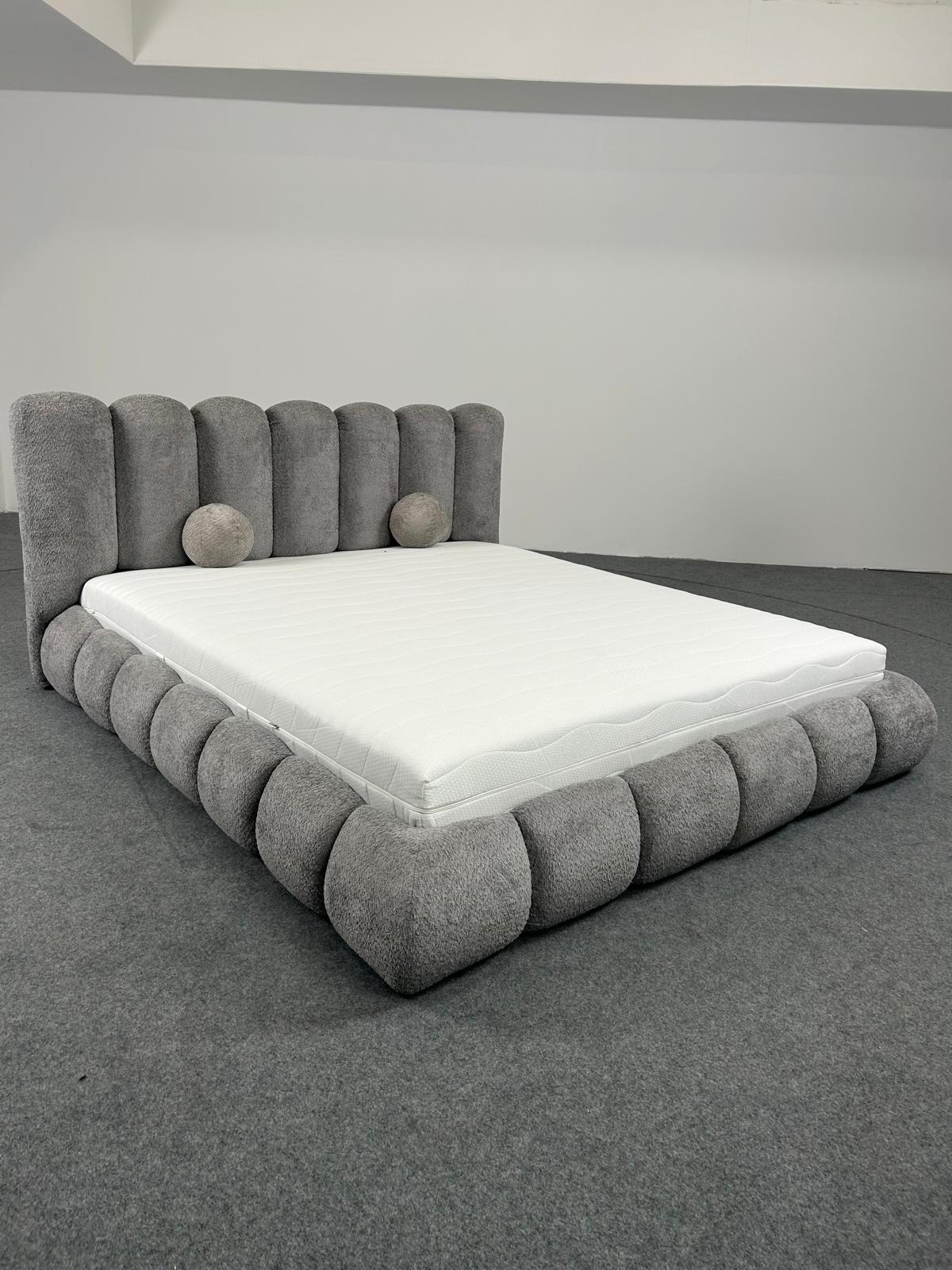 Szare, tapicerowane łóżko z okrągłymi elementami i materacem, sfotografowane w studiu na szarym tle. Widok z przodu, lekko z boku, ukazujący unikalny design.
