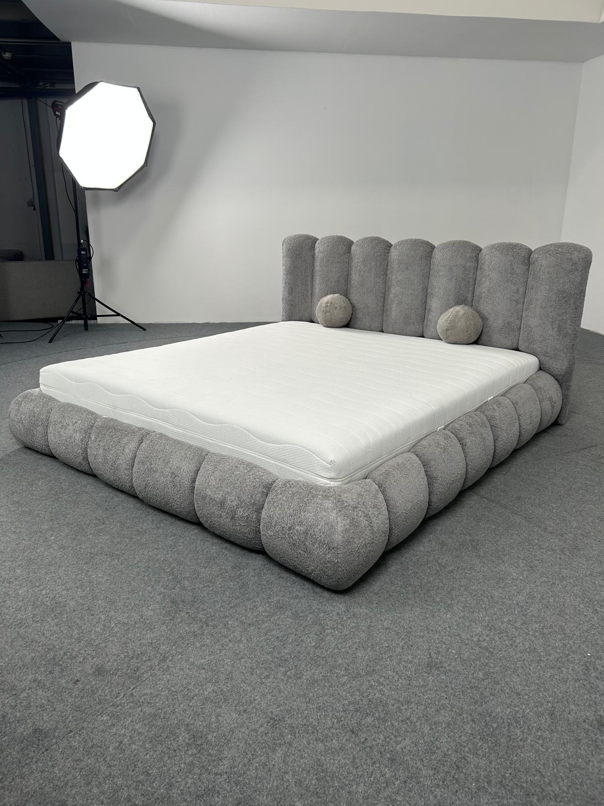 Szare, tapicerowane łóżko z okrągłymi poduszkami i materacem na szarym dywanie, oświetlone lampą studyjną. Nietypowy design.