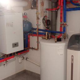 R.B INSTAL Rafał Bajszczak - Biały piec gazowy z podłączeniami rur w kolorze niebieskim i czerwonym, zbiornik na wodę i zmiękczacz wody Keller w kotłowni. Instalacja po montażu.