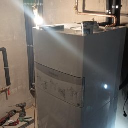 R.B INSTAL Rafał Bajszczak - Biały piec gazowy Vaillant w piwnicy, z widocznymi rurami miedzianymi i narzędziami obok. Oświetlenie sufitowe tworzy refleksy na obudowie pieca.