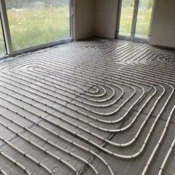 R.B INSTAL Rafał Bajszczak - Białe rury ogrzewania podłogowego ułożone spiralnie na szarej folii w jasnym pomieszczeniu z dużymi oknami, widok na zewnątrz na zieleń.