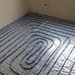 R.B INSTAL Rafał Bajszczak - Niebieskie rury ogrzewania podłogowego ułożone na folii z siatką w surowym wnętrzu, widoczne okno i ściany przed wylewką. Instalacja Aque Team.
