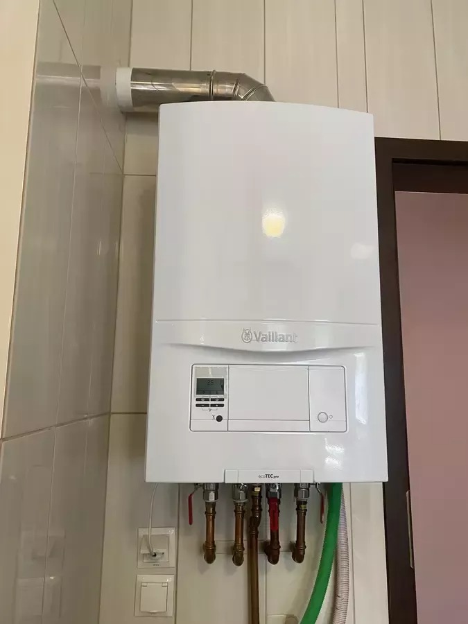 Biały, wiszący piec gazowy Vaillant z podłączonymi rurami miedzianymi i zielonym wężem, zamontowany na ścianie z jasnymi płytkami. Widoczny fragment rury odprowadzającej spaliny.