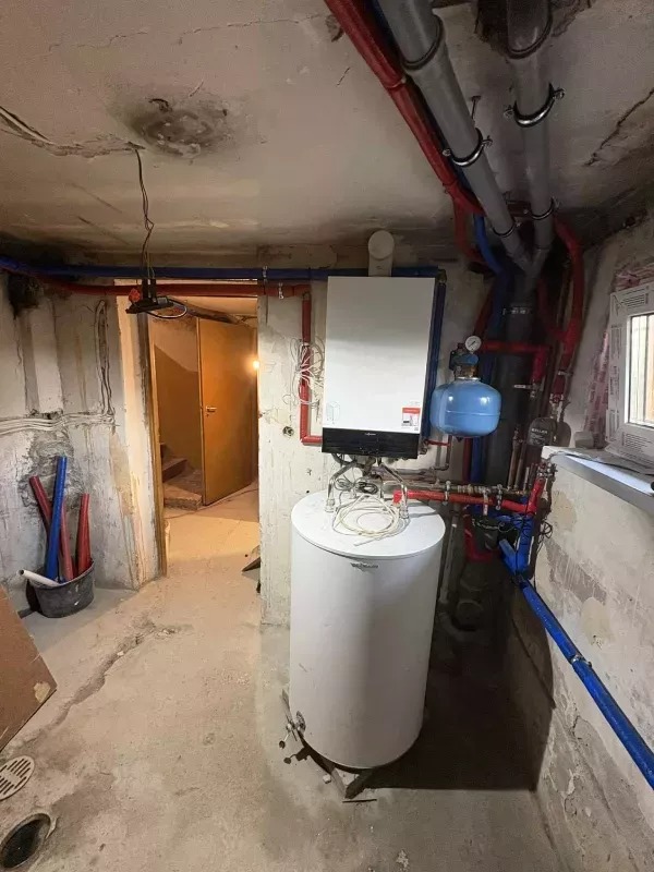 Biały bojler i piec gazowy na tle surowego wnętrza piwnicy z widocznymi rurami instalacji CO i wodnej. Otwór drzwiowy w tle.