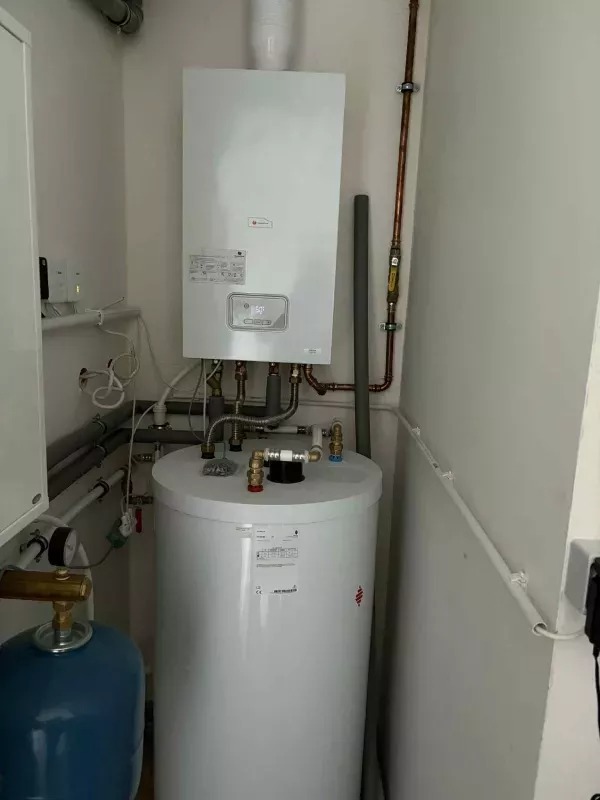 Biały, wiszący piec gazowy z podgrzewaczem wody w kotłowni. Widoczne rury miedziane i plastikowe. Na ścianie zamontowane urządzenia sterujące.
