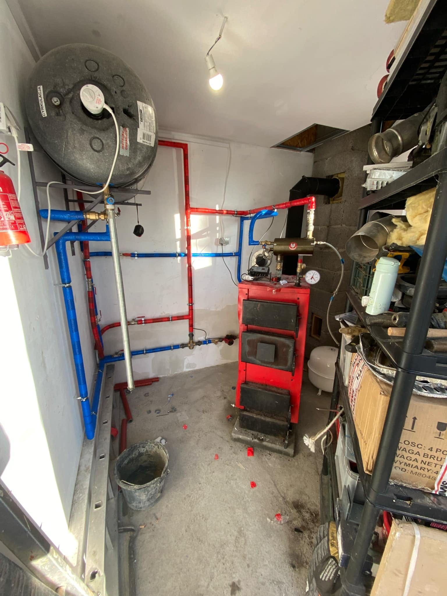 Czerwony piec na paliwo stałe z podłączonymi rurami (czerwone i niebieskie) w małym pomieszczeniu. Widoczny zbiornik na wodę, drabina i regał z narzędziami.