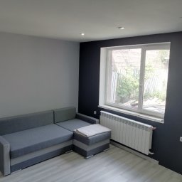 Łukasz Borycki - Szaro-grafitowe wnętrze pokoju po odświeżeniu: narożnik, okno z grzejnikiem, punktowe oświetlenie sufitowe. Kontrastowe ściany i jasna podłoga.