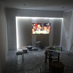 Łukasz Borycki - Pokój w trakcie prac wykończeniowych: ściany przygotowane do malowania, zabezpieczona folią podłoga, drabina, oświetlenie LED wokół telewizora. Widoczne narzędzia i materiały.