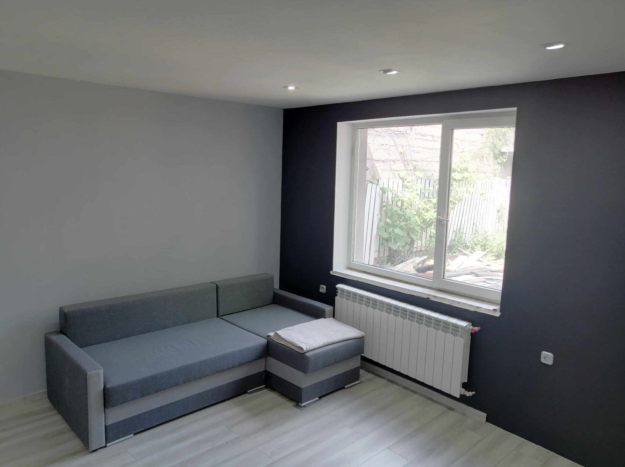 Szaro-grafitowe wnętrze pokoju po odświeżeniu: narożnik, okno z grzejnikiem, punktowe oświetlenie sufitowe. Kontrastowe ściany i jasna podłoga.