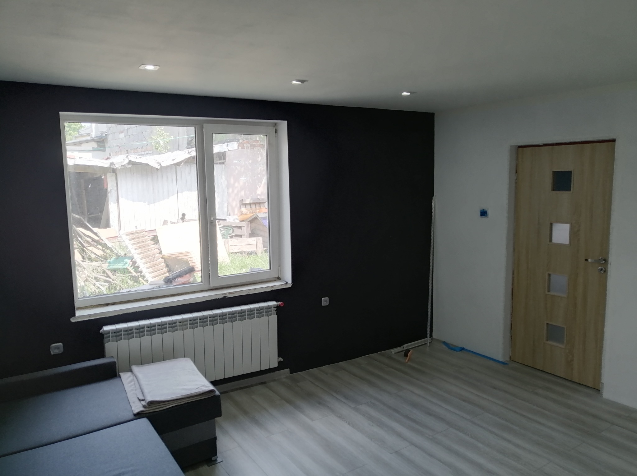 Pokój po remoncie: ciemna ściana, jasne panele podłogowe, biały grzejnik pod oknem. Widoczny fragment sofy i drzwi. Minimalistyczny wystrój.