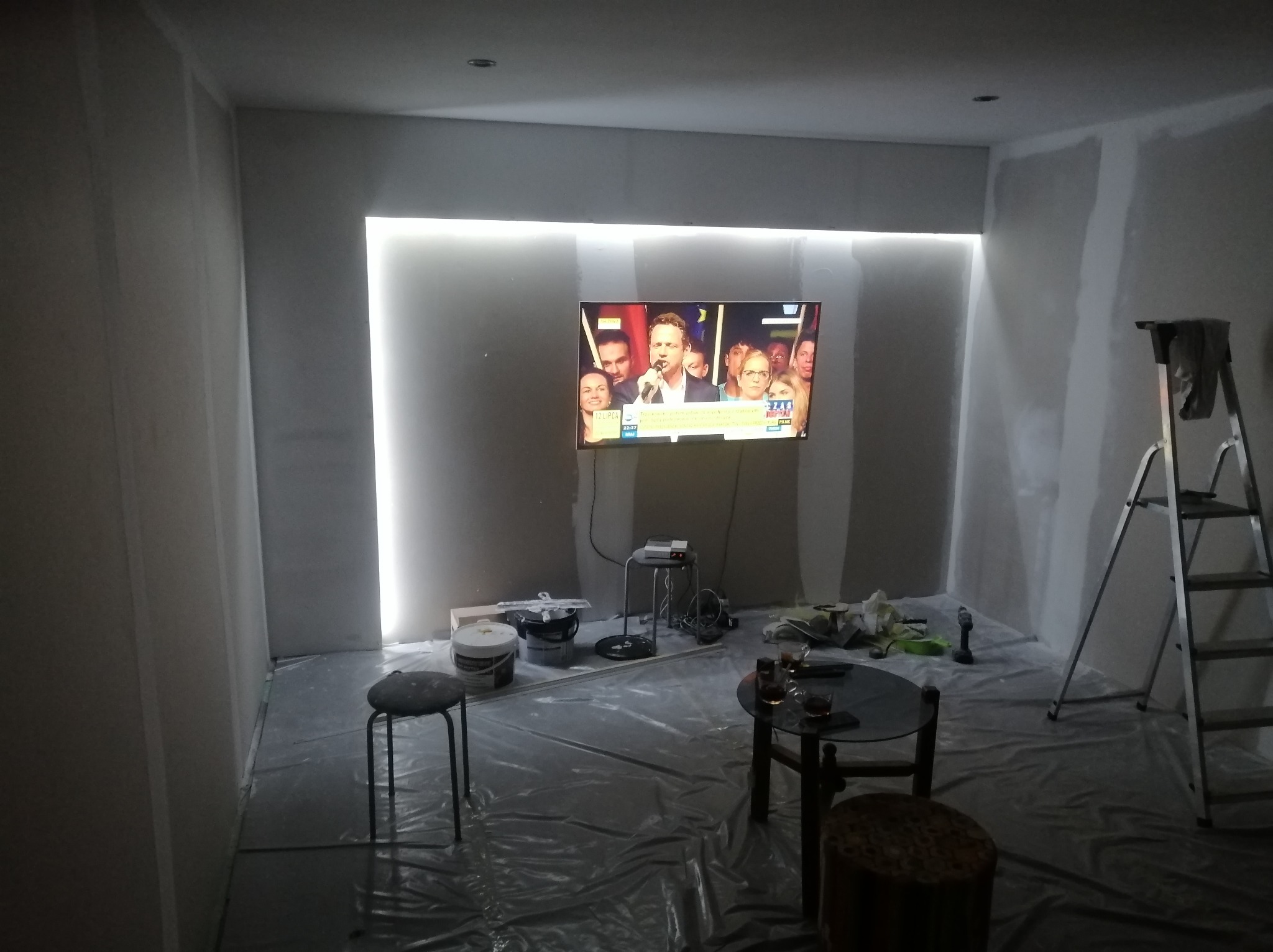 Pokój w trakcie prac wykończeniowych: ściany przygotowane do malowania, zabezpieczona folią podłoga, drabina, oświetlenie LED wokół telewizora. Widoczne narzędzia i materiały.