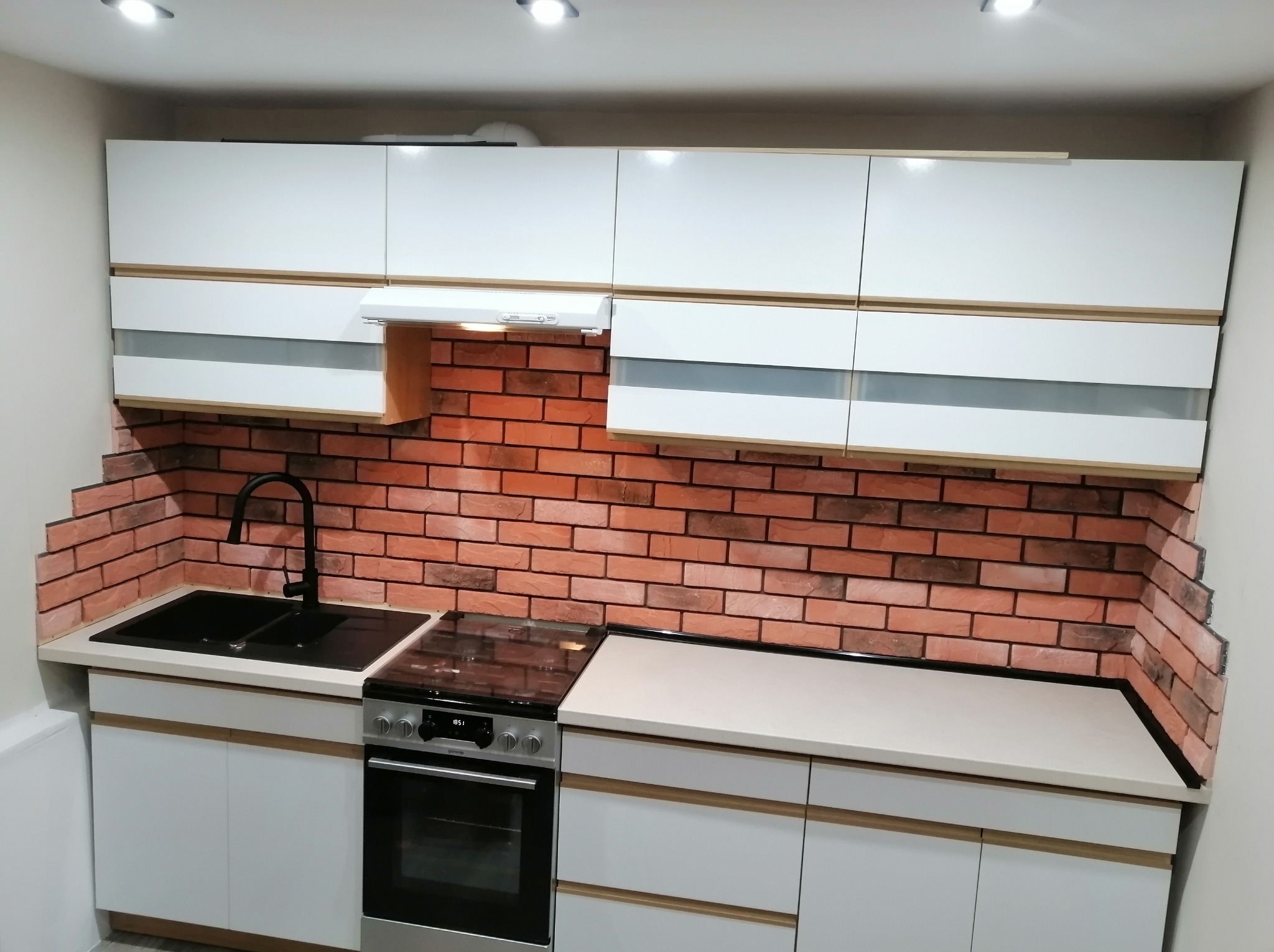 Nowoczesna kuchnia z białymi szafkami i ceglaną ścianą. Czarny zlew i piekarnik. Jasne oświetlenie punktowe. Minimalistyczny design z akcentem rustykalnym.