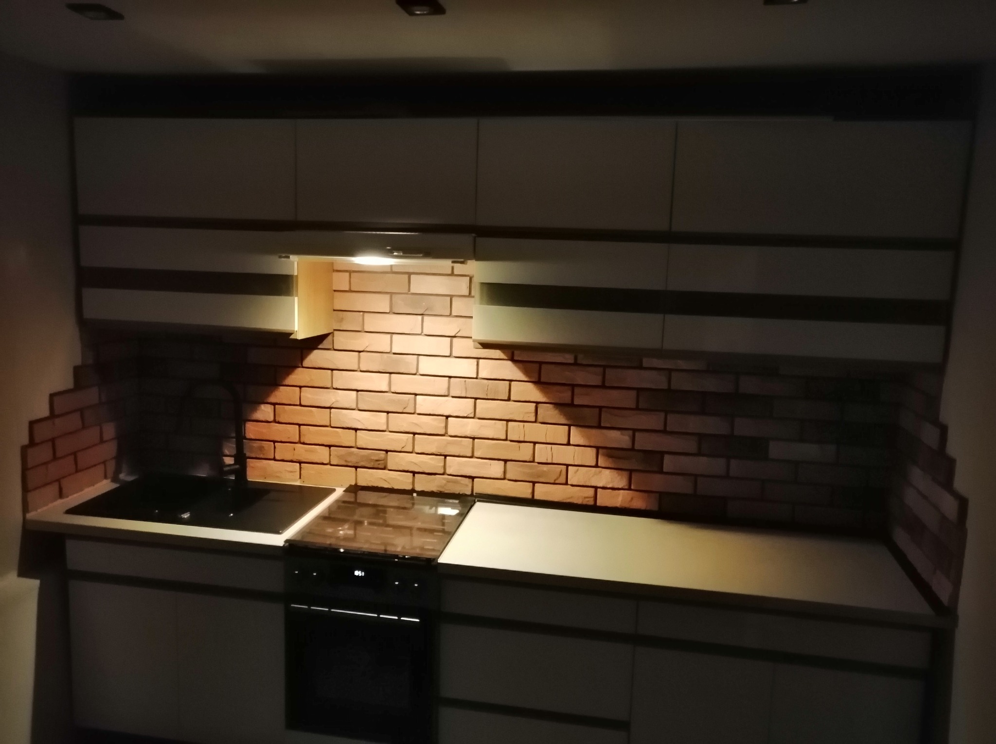 Nowoczesna kuchnia z ceglaną ścianą nad blatem, oświetlona punktowo, z ciemnymi szafkami dolnymi i jasnymi górnymi. Widok z góry.