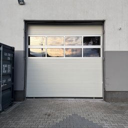 F.H.U. RUPMET ŁUKASZ MADETKO - Biała segmentowa brama garażowa z oknami, odbijającymi zachód słońca, zamontowana w szarej elewacji budynku. Widoczna kostka brukowa i kontener.