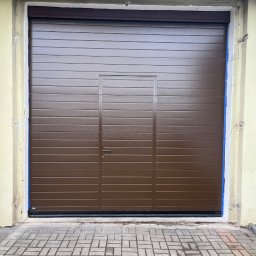 F.H.U. RUPMET ŁUKASZ MADETKO - Zamknięta, brązowa brama garażowa segmentowa z drzwiami wejściowymi pośrodku, widoczna z zewnątrz na tle betonowej kostki brukowej i fragmentu elewacji budynku.