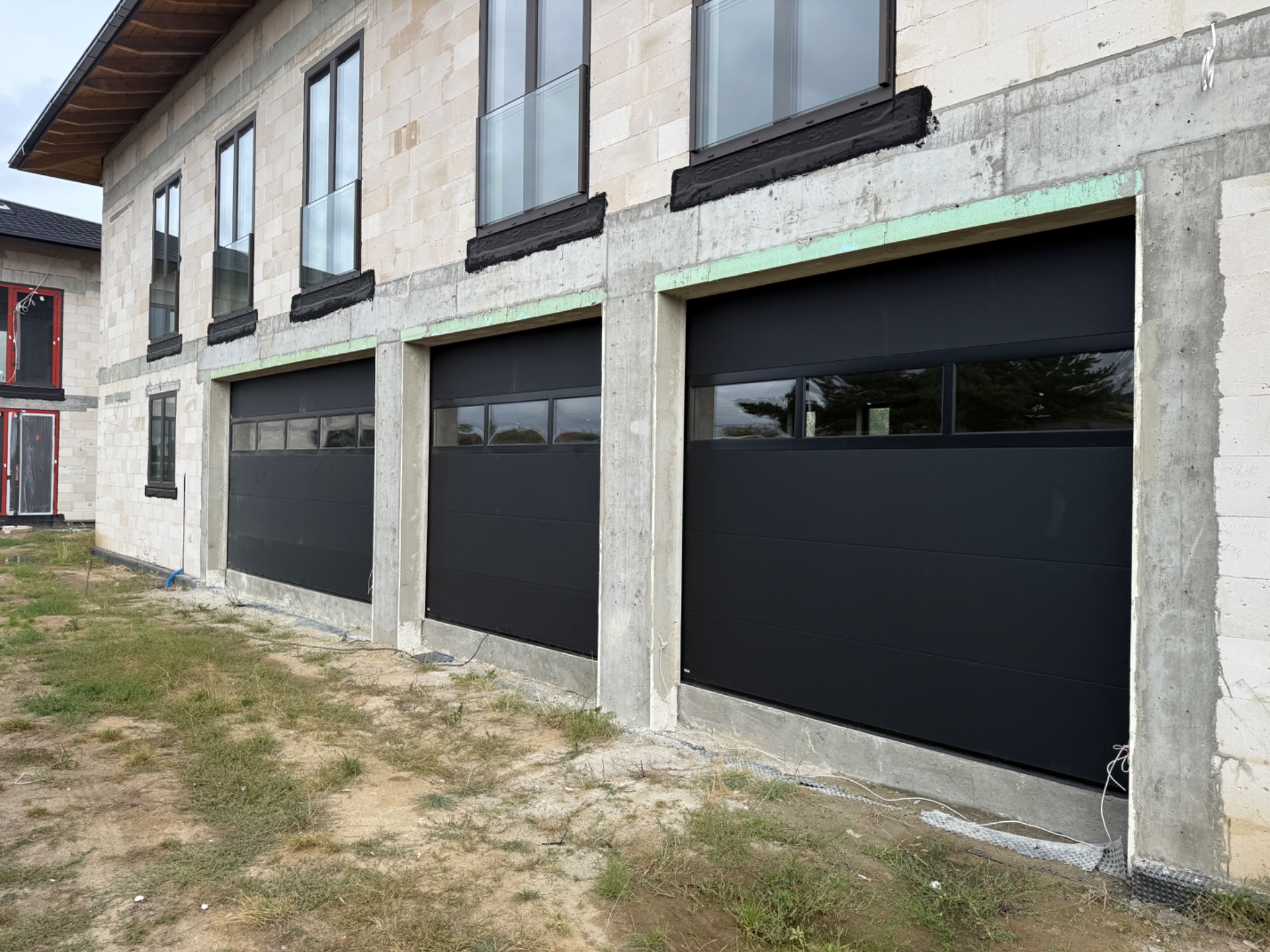 Nowoczesny budynek w Pęcicach z trzema czarnymi bramami garażowymi segmentowymi, widoczne okna na piętrze i surowy stan elewacji.