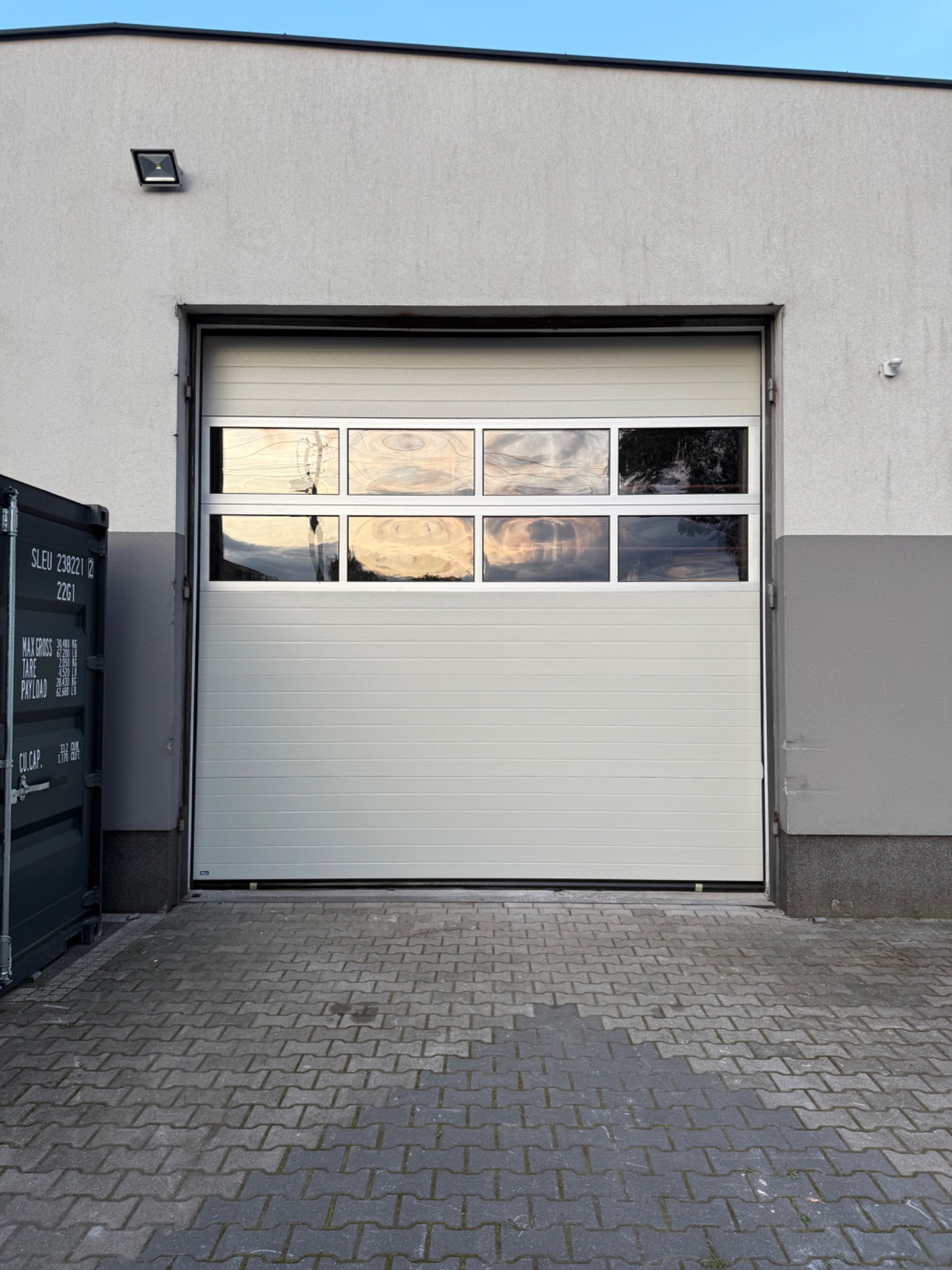 Biała segmentowa brama garażowa z oknami, odbijającymi zachód słońca, zamontowana w szarej elewacji budynku. Widoczna kostka brukowa i kontener.