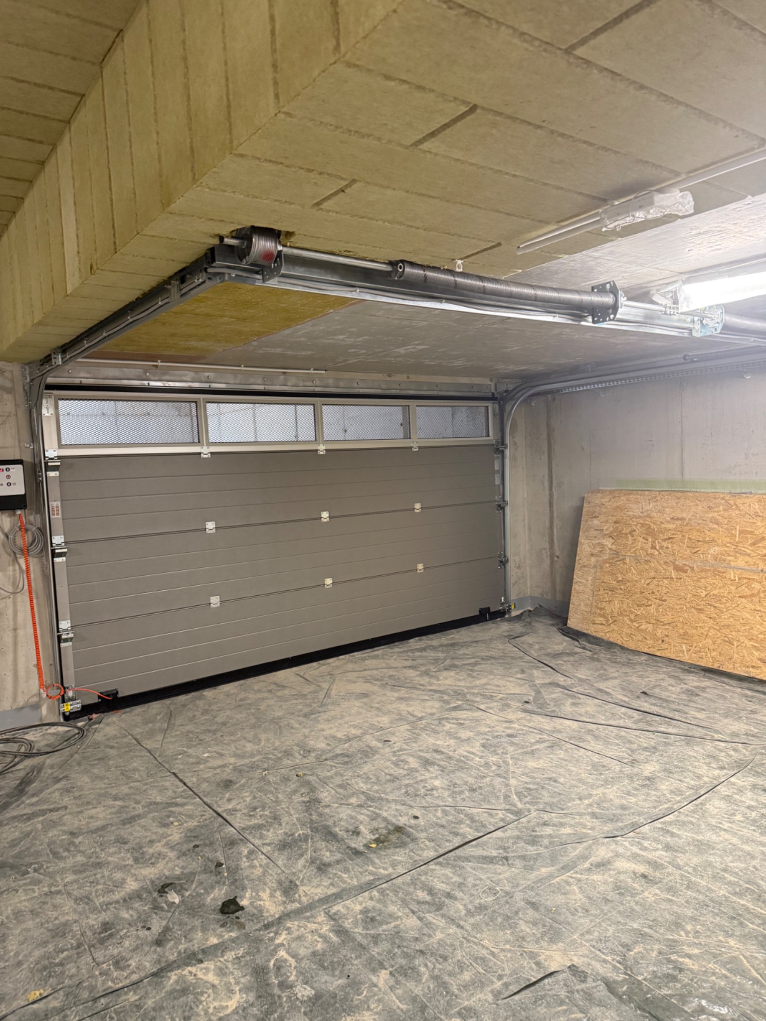Szara segmentowa brama garażowa z oknami u góry, widoczny mechanizm otwierania pod sufitem, zabezpieczona folią podłoga, płyta OSB oparta o ścianę.