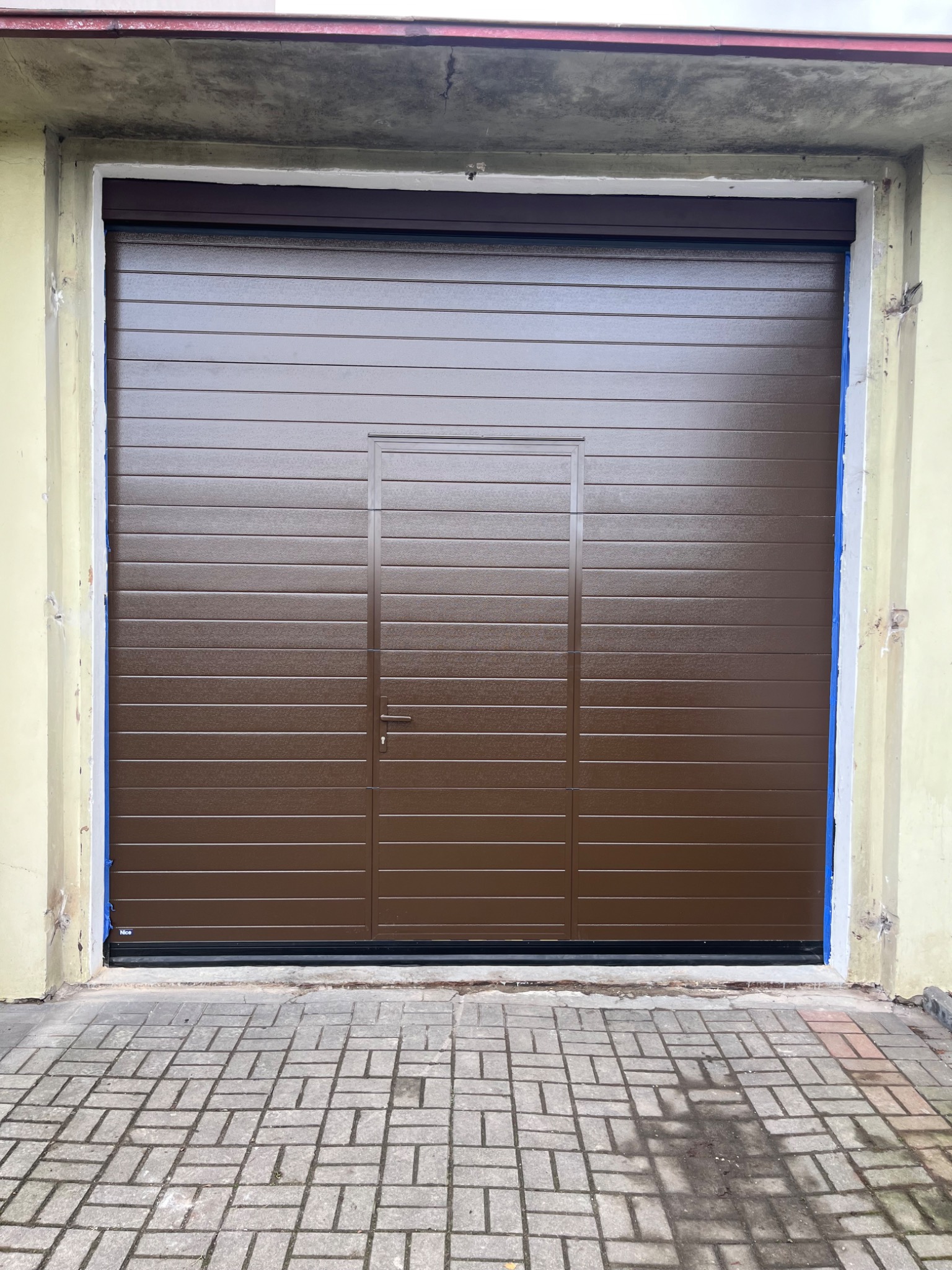Zamknięta, brązowa brama garażowa segmentowa z drzwiami wejściowymi pośrodku, widoczna z zewnątrz na tle betonowej kostki brukowej i fragmentu elewacji budynku.