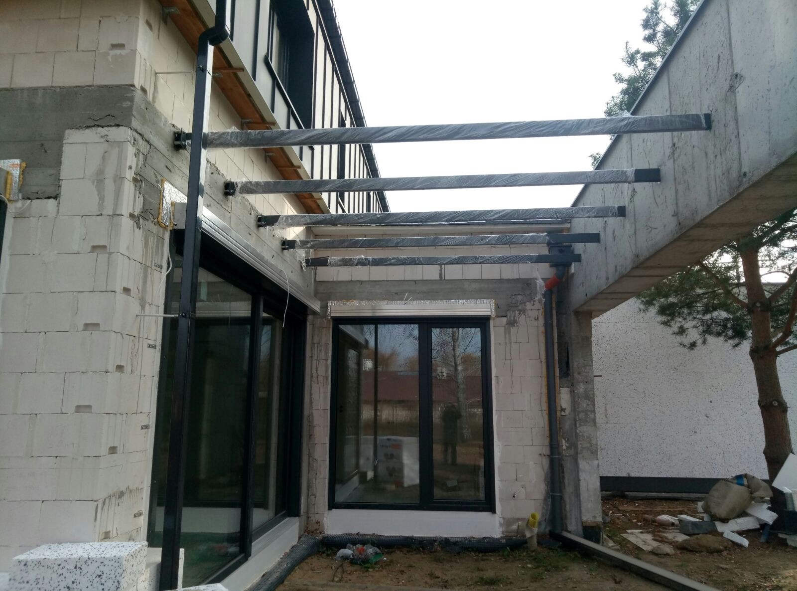 Metalowa pergola w trakcie montażu, zabezpieczona folią, przymocowana do surowej ściany budynku z oknami. Widoczne elementy konstrukcyjne i materiały budowlane.