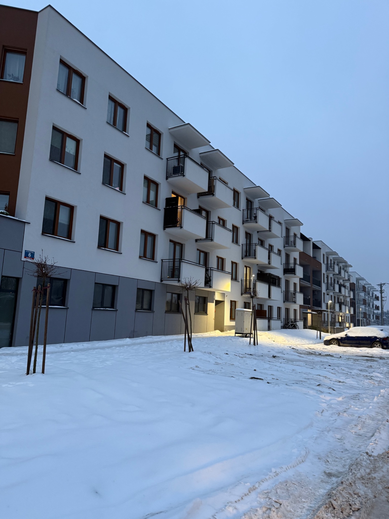 Nowoczesny budynek wielorodzinny z balkonami i czarnymi balustradami, ośnieżony teren, pochmurne niebo. Widok na elewację z numerem 6.