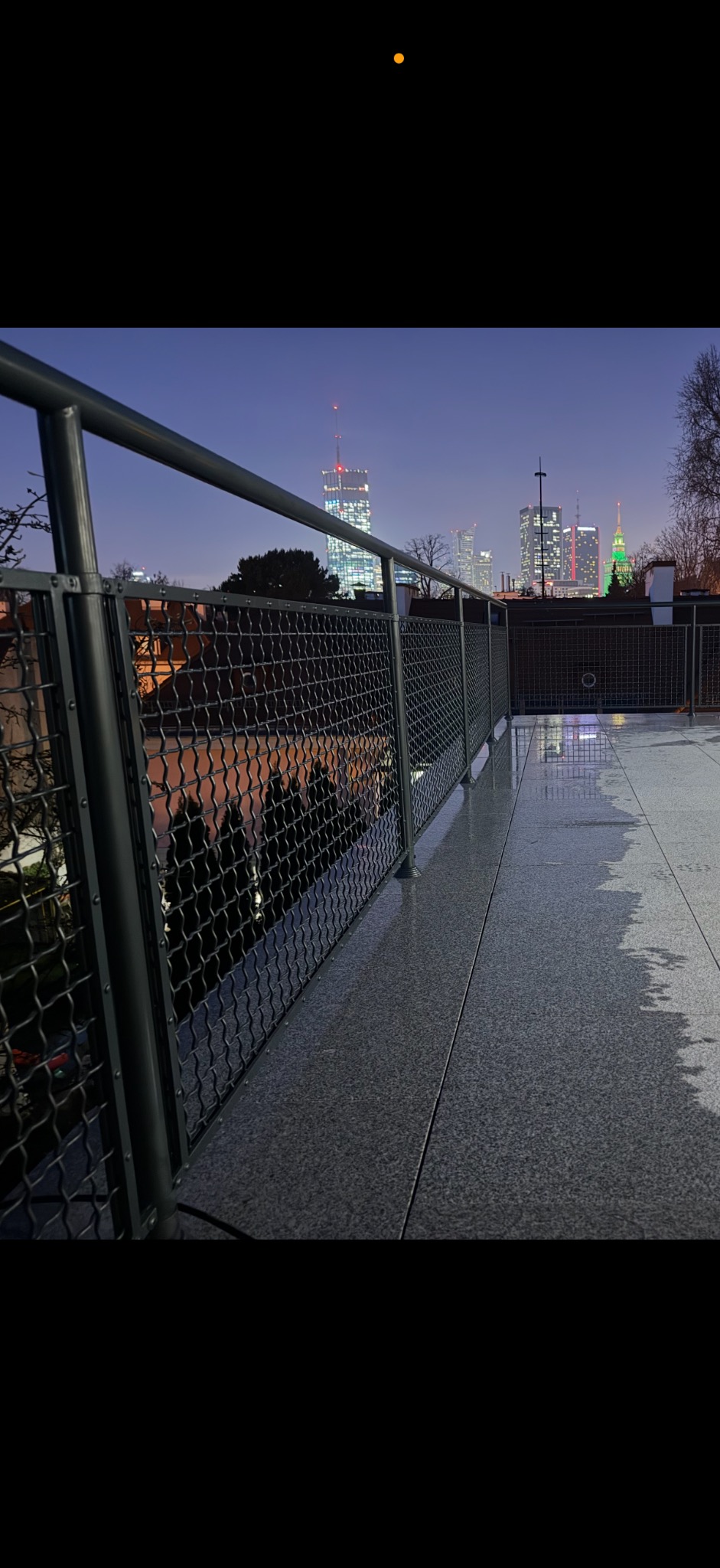Metalowa balustrada balkonowa z widokiem na panoramę miasta nocą. Szara, mokra nawierzchnia balkonu odbija światła miejskie.